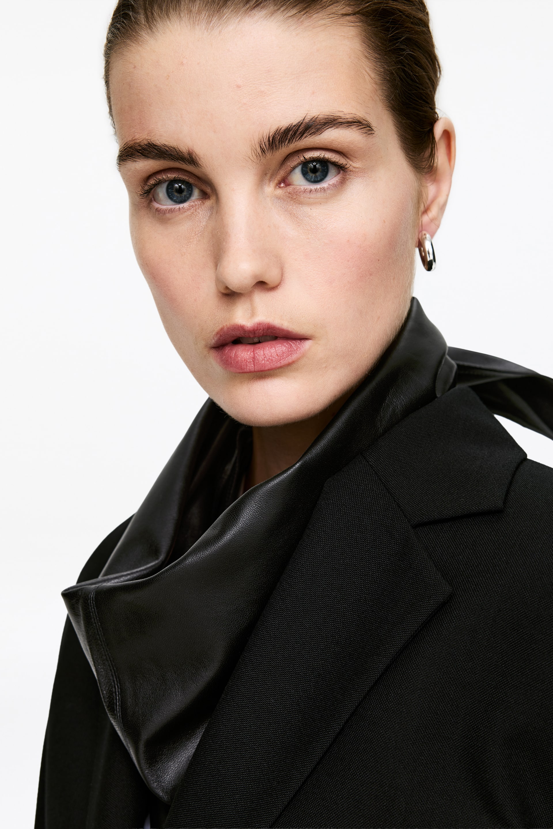 View larger image: A black blazer is being presented from a slight front-side angle, showcasing a structured lapel. A black, smooth, leather-like element is draped around the neck and shoulder area, creating a cowl-like layer over the blazer.
