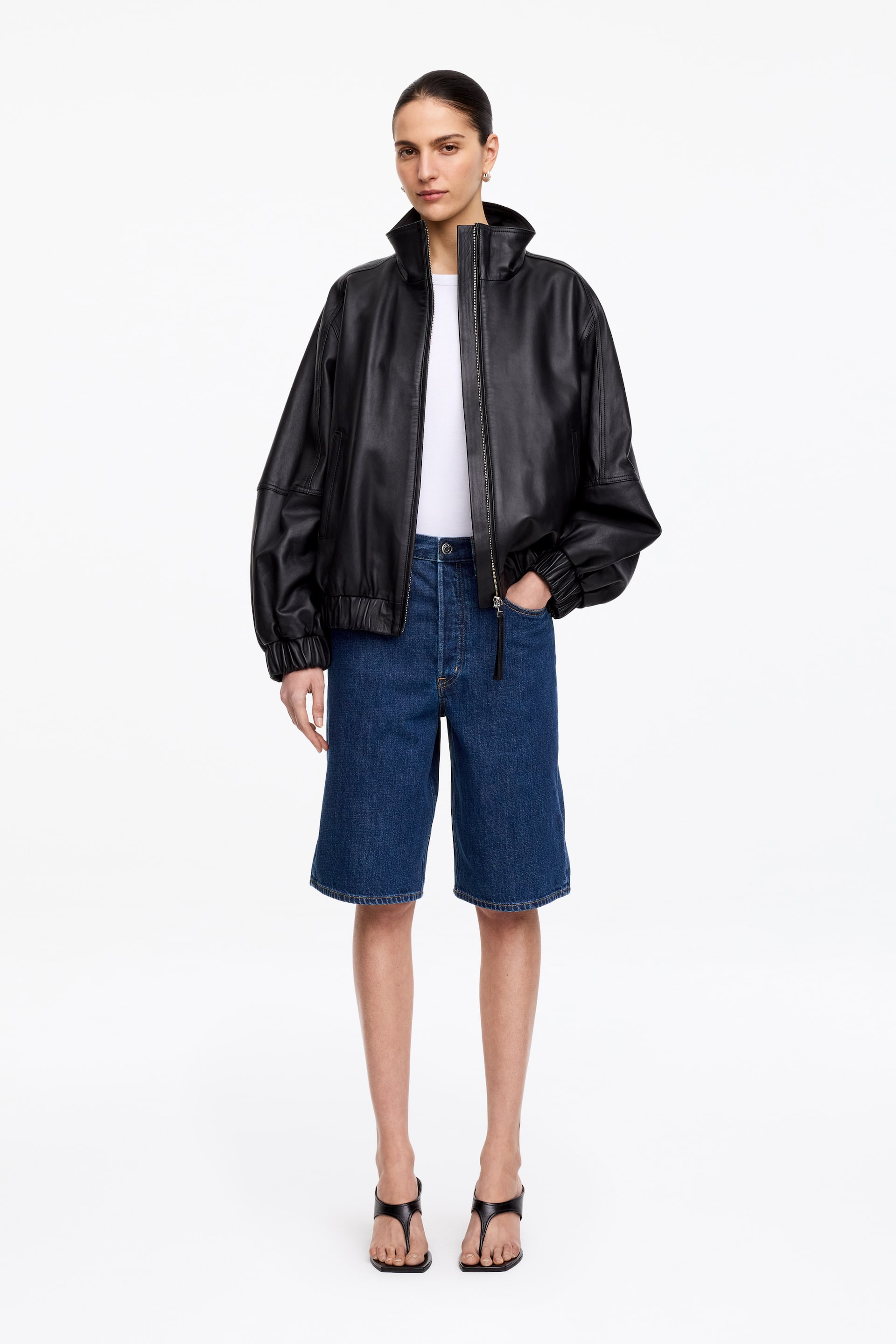 Vezi imaginea mai mare: Jacheta bomber din piele neagră prezintă un croi supradimensionat, un guler înalt și fermoar frontal argintiu, parțial deschis. Este proiectată cu mâneci lungi și pline, cu manșete elastice adunate și tiv elasticizat, fiind purtată peste un tricou alb.