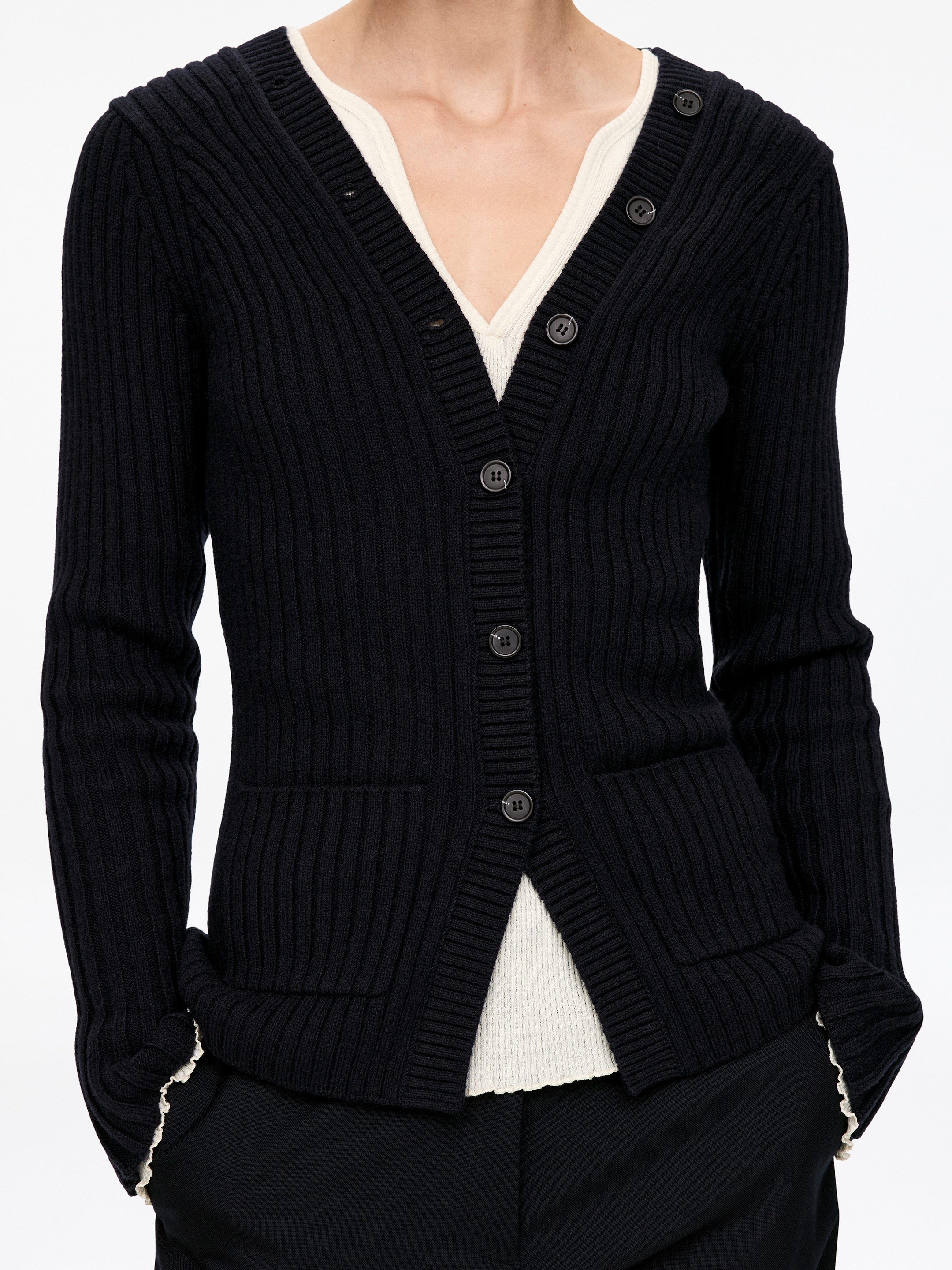 Visualizza immagine più grande: Un cardigan nero a maglia a coste presenta uno scollo a V, una chiusura con bottoni sul davanti con bottoni scuri, maniche lunghe e due tasche applicate frontali. Sotto è stratificato un top a coste color panna, con lo scollo, l'orlo e i polsini visibili.