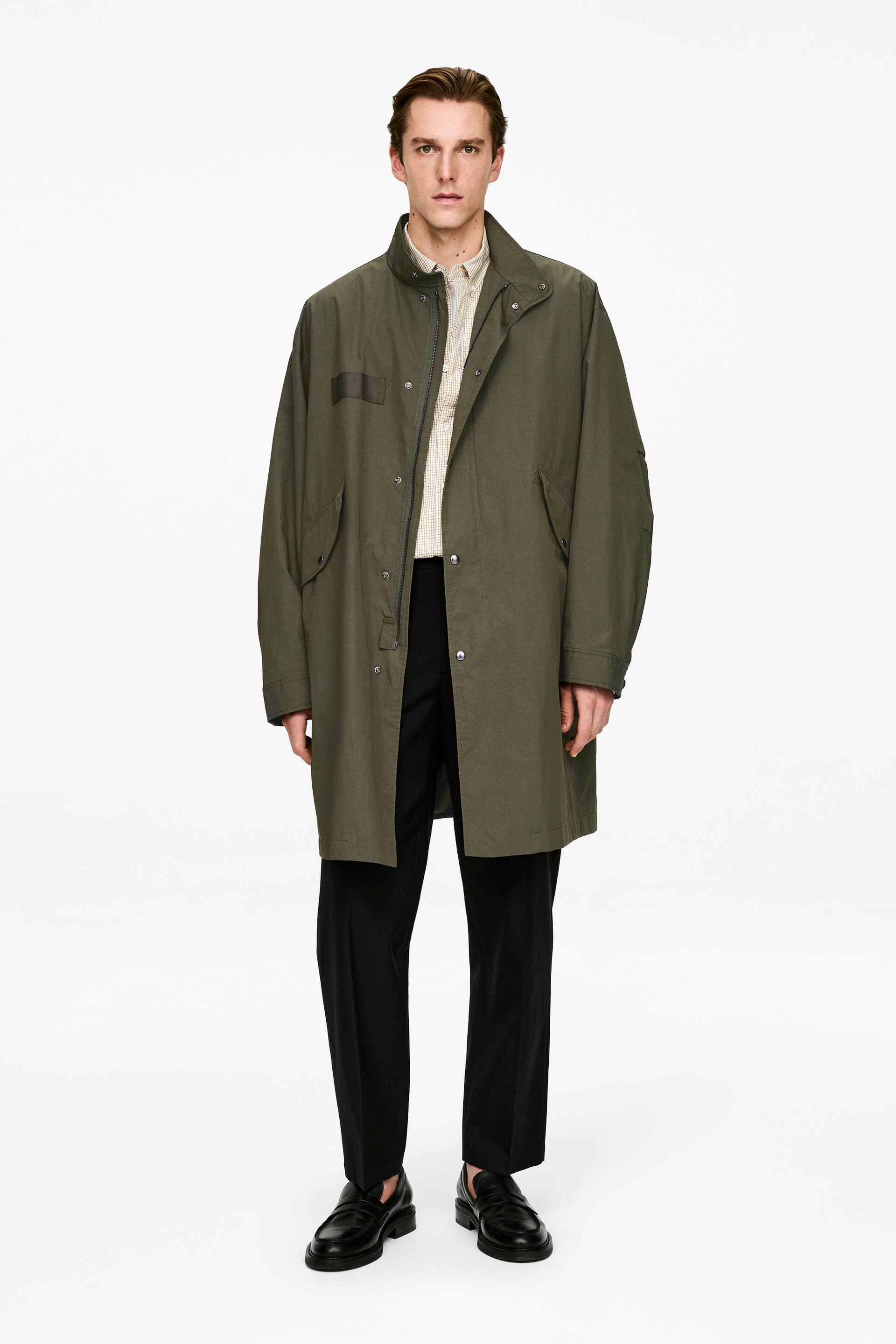 Προβολή μεγαλύτερης εικόνας: Ένα παλτό σε λαδί χρώμα παρουσιάζεται από μπροστά, διαθέτοντας μια χαλαρή σιλουέτα μέχρι το μέσο του μηρού και έναν όρθιο γιακά. Διαθέτει μερικό μπροστινό κλείσιμο με φερμουάρ που καλύπτεται από μια πατιλέτα με κουμπιά σούστες, δύο μεγάλες τσέπες με καπάκι στο κάτω μπροστινό μέρος και μια ορθογώνια λεπτομέρεια με έμπλαστρο στο πάνω αριστερό στήθος, με ένα ελαφρύ καρό πουκάμισο να είναι ορατό από κάτω.