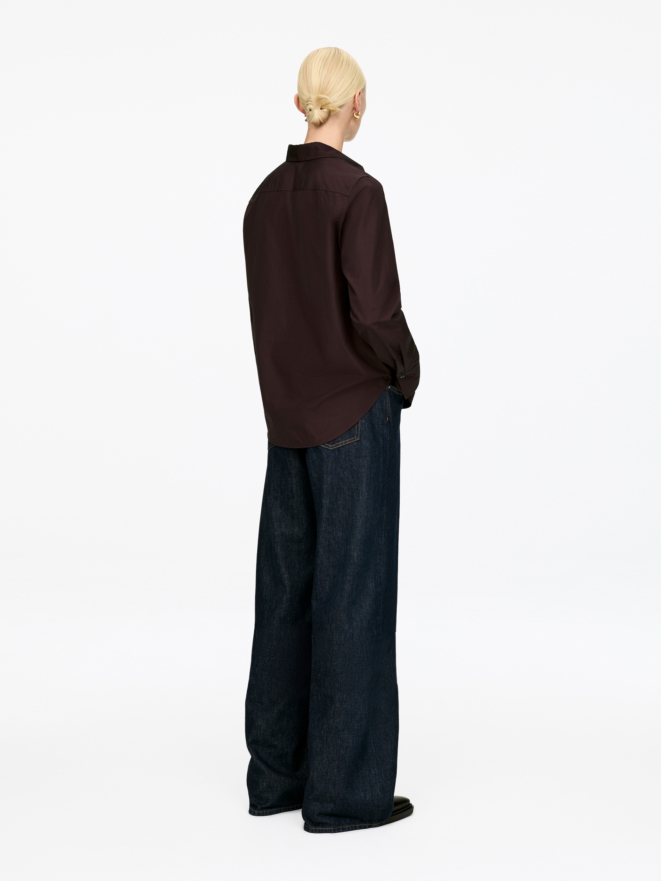 Ingrandisci l'immagine: Una camicia con bottoni a maniche lunghe di colore marrone scuro è presentata con una vista posteriore, caratterizzata da una vestibilità rilassata e parzialmente infilata in vita. È indossata con jeans blu scuro a gamba larga che mostrano un taglio lungo e ampio.