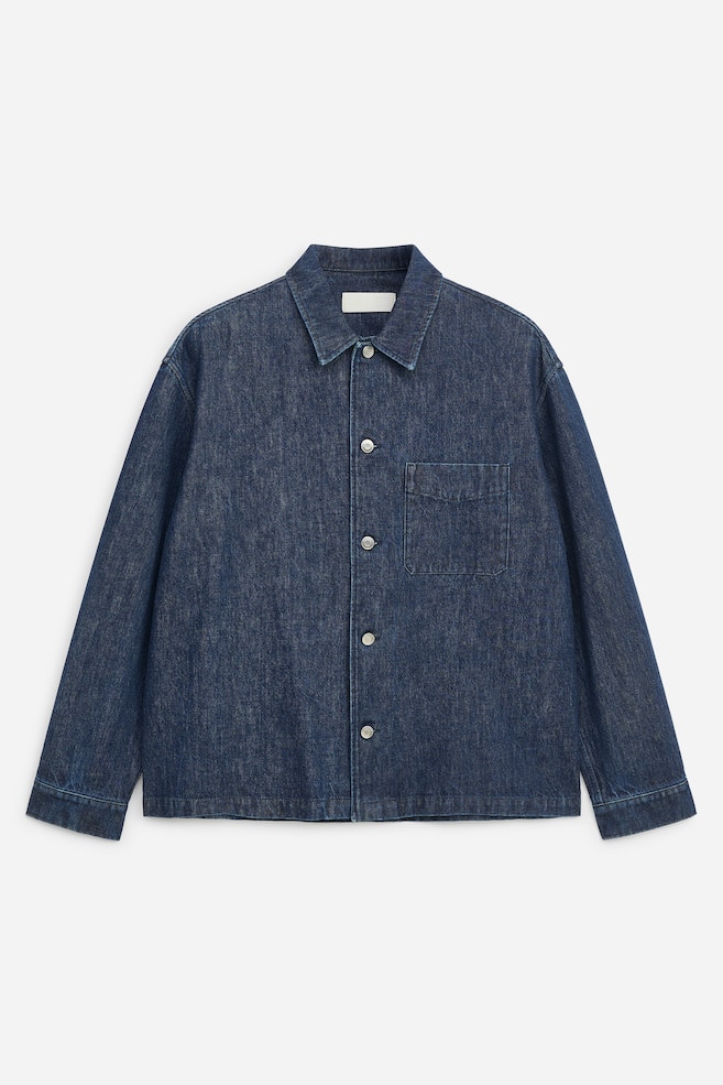 A dark blue denim jacket is shown from the front, featuring a point collar, a silver-tone button-up front fastening, and a single patch pocket on the chest. The jacket has long sleeves with simple cuffs and a straight hem, giving it a relaxed fit. - 1