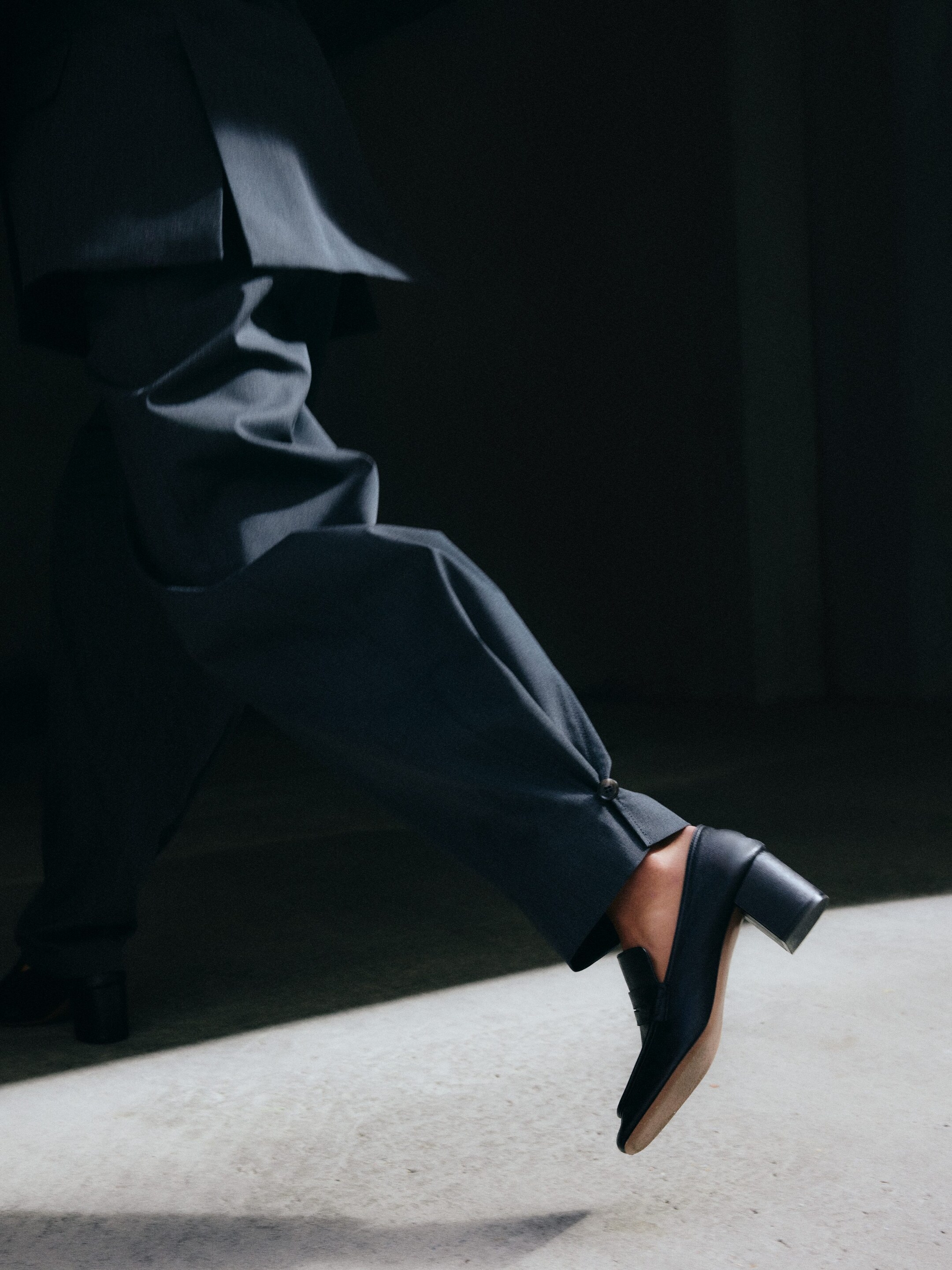 Größeres Bild anzeigen: Ein schwarzer Loafer-Schuh aus Leder mit Blockabsatz und hellfarbener Sohle wird getragen und ist nach links geneigt. Dunkelgraue Hosen mit Knöchelknopf fallen über den Schuh.