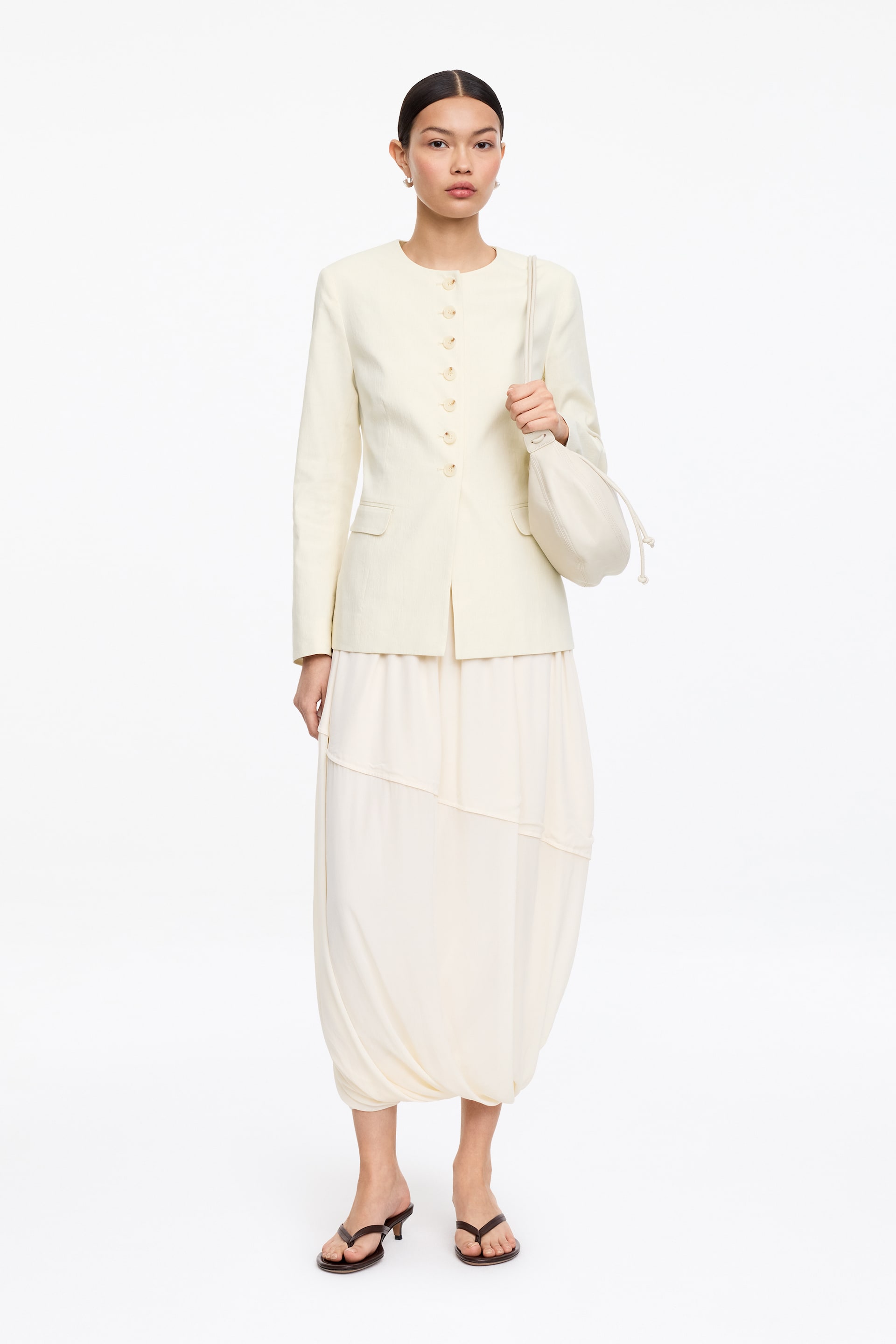 Ver imagen más grande: Una chaqueta crema sin cuello presenta un cierre de seis botones y dos bolsillos de parche, mientras cubre una falda globo completa en crema que cuenta con una cintura fruncida y paneles asimétricos. Se sostiene un bolso de mano con cordón a juego en crema, y unas sandalias de dedo marrón oscuro con tacones bajos completan el conjunto.
