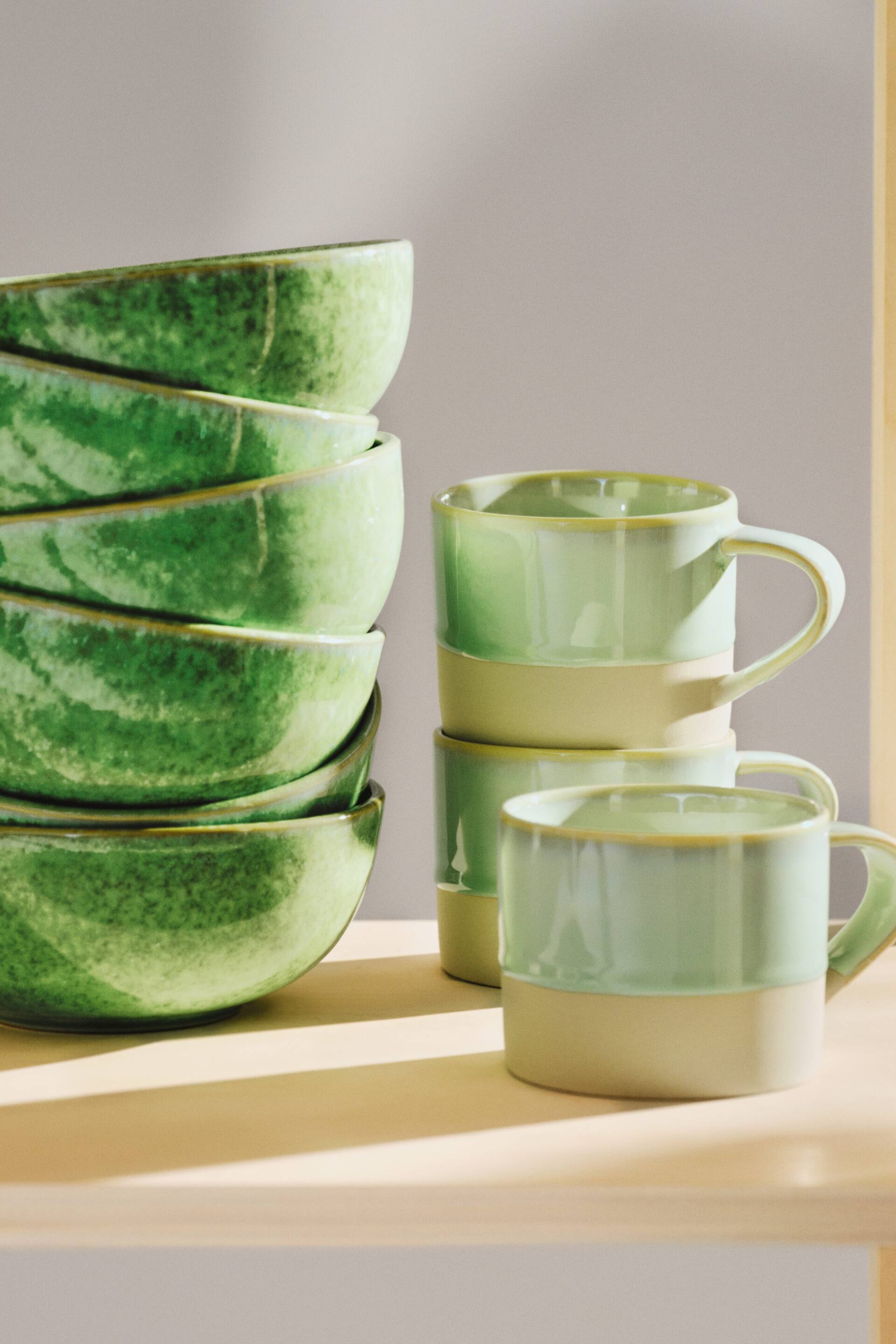 Agrandir l'image: Cinq bols en céramique verts sont empilés sur la gauche, légèrement inclinés vers l'avant, affichant un émail moucheté sur leurs surfaces et intérieurs. Trois tasses en céramique bicolores sont empilées sur la droite, légèrement inclinées vers la droite, avec une section supérieure vert clair, une section inférieure crème, et des anses visibles.