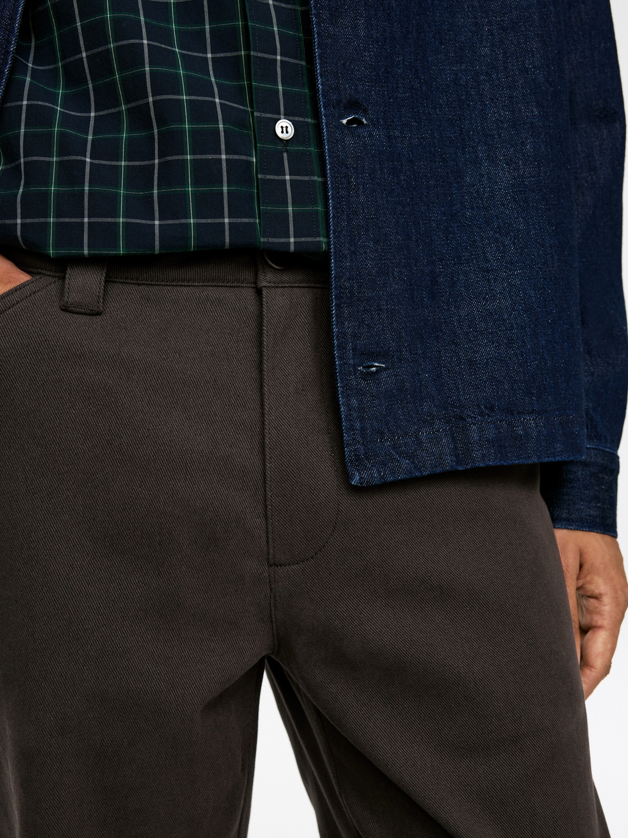 Visualizza immagine più grande: Pantaloni marrone scuro, caratterizzati da una distintiva trama a spina di pesce, sono esposti dalla parte superiore della coscia e dell'anca. I pantaloni presentano passanti per cintura e una cucitura frontale curva, con una mano inserita nella tasca laterale sinistra. Una camicia a quadri è indossata infilata nei pantaloni, stratificata sotto una giacca di jeans scura.