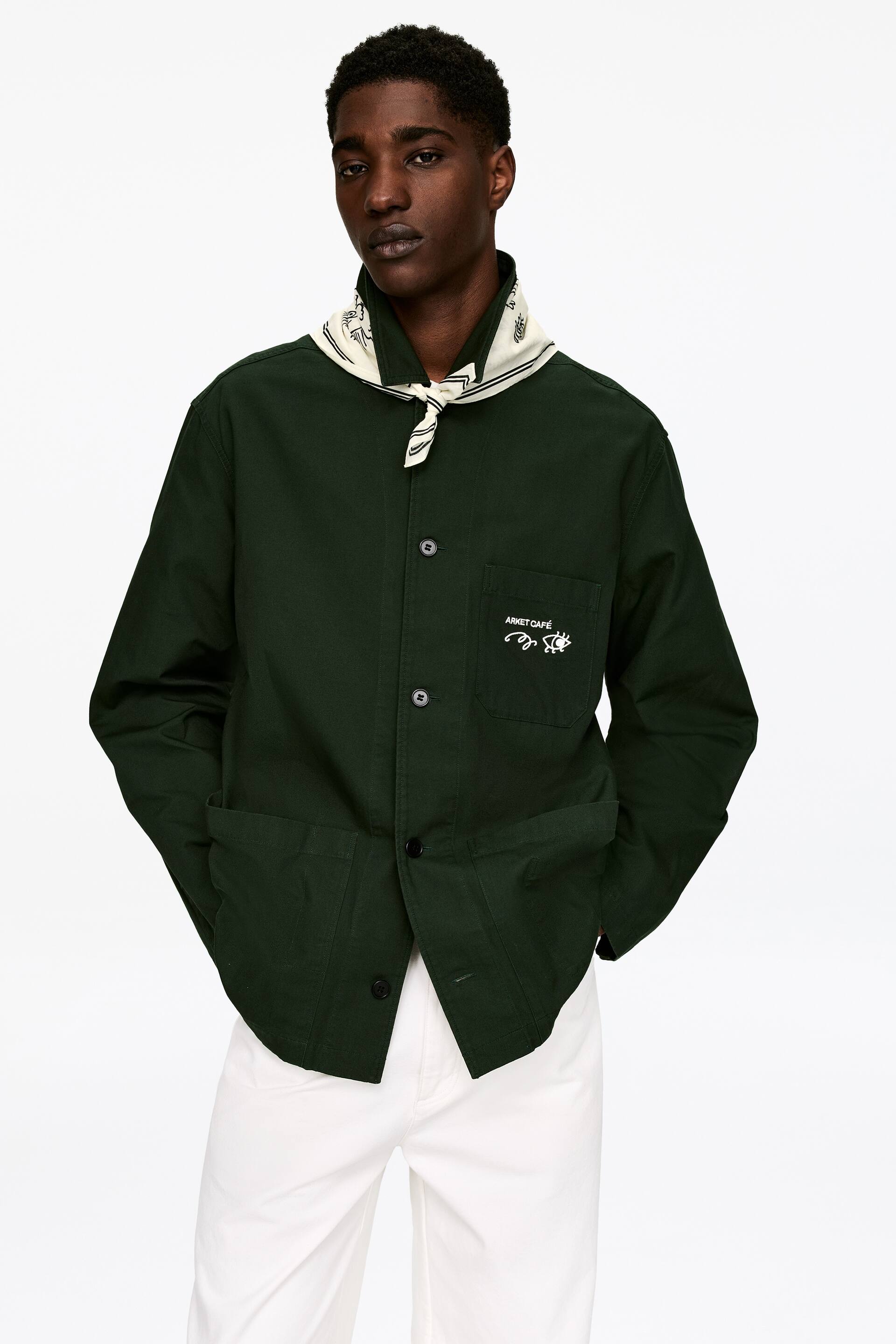 Vis større bilde: En dyp grønn arbeidsjakke vises fra forsiden, med en spiss krage, en knappelukking foran med mørke knapper og lange ermer. Den har en brystlomme med "ARKET CAFÉ" brodert i hvitt og to nedre påsydde lommer, og er stylet med et kremfarget bandana knyttet rundt halsen over kragen.