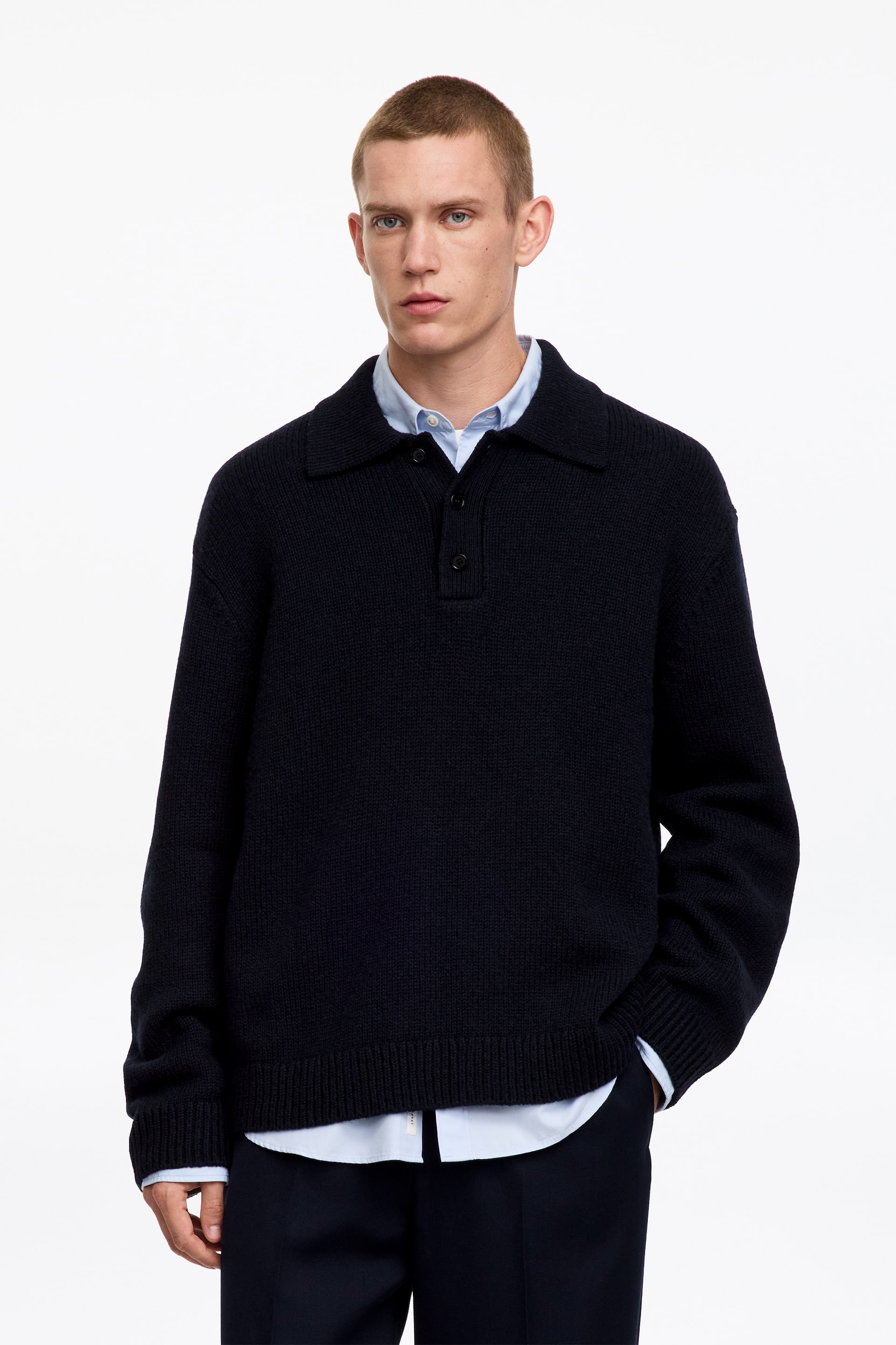 Agrandir l'image: Un pull polo en maille bleu marine foncé, avec col polo et patte de boutonnage à mi-longueur, est porté par le mannequin. Sa coupe décontractée est superposée à une chemise bleu clair à col, visible au niveau du cou et de l’ourlet.