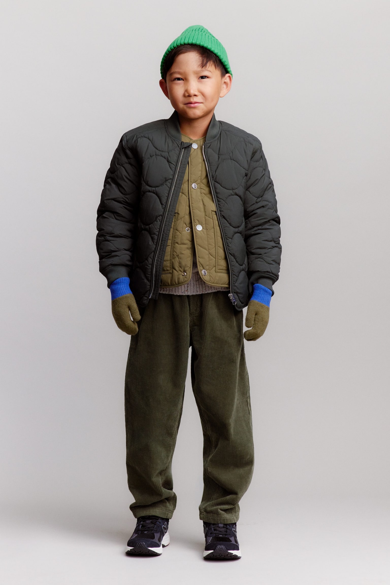 Ein Kind steht frontal und trÃ¤gt eine leuchtend grÃ¼ne, gerippte BaumwollmÃ¼tze mit umgeschlagenem Saum, eine dunkelgraue, leicht gesteppte Jacke mit Zwiebelsteppung, offen getragen Ã¼ber einer olivgrÃ¼nen, gesteppten Weste mit Druckknopfverschluss, sowie locker geschnittene, olivgrÃ¼ne Cordhosen. Abgerundet wird der Look durch olivgrÃ¼ne WollfÃ¤ustlinge mit leuchtend blauen, gerippten BÃ¼ndchen und dunkelgraue New Balance Trainers mit weissen Sohlen. - 1