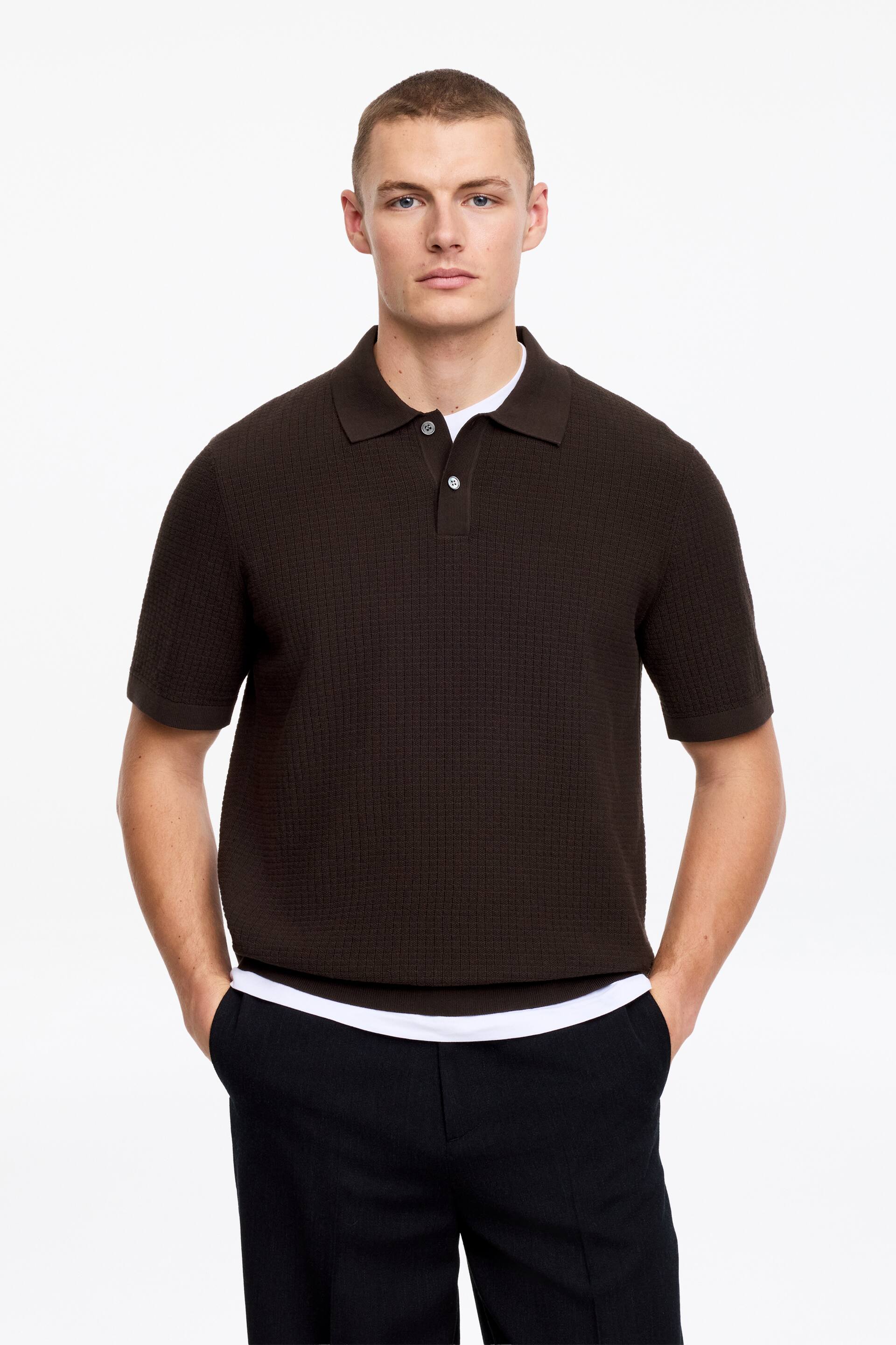 Größeres Bild ansehen: Ein braunes Poloshirt wird von vorne gezeigt, das ein strukturiertes Strickmuster, eine Zwei-Knopf-Leiste mit hellen Knöpfen und kurze Ärmel aufweist. Ein weißes Unterhemd ist unter dem gerippten Saum sichtbar.