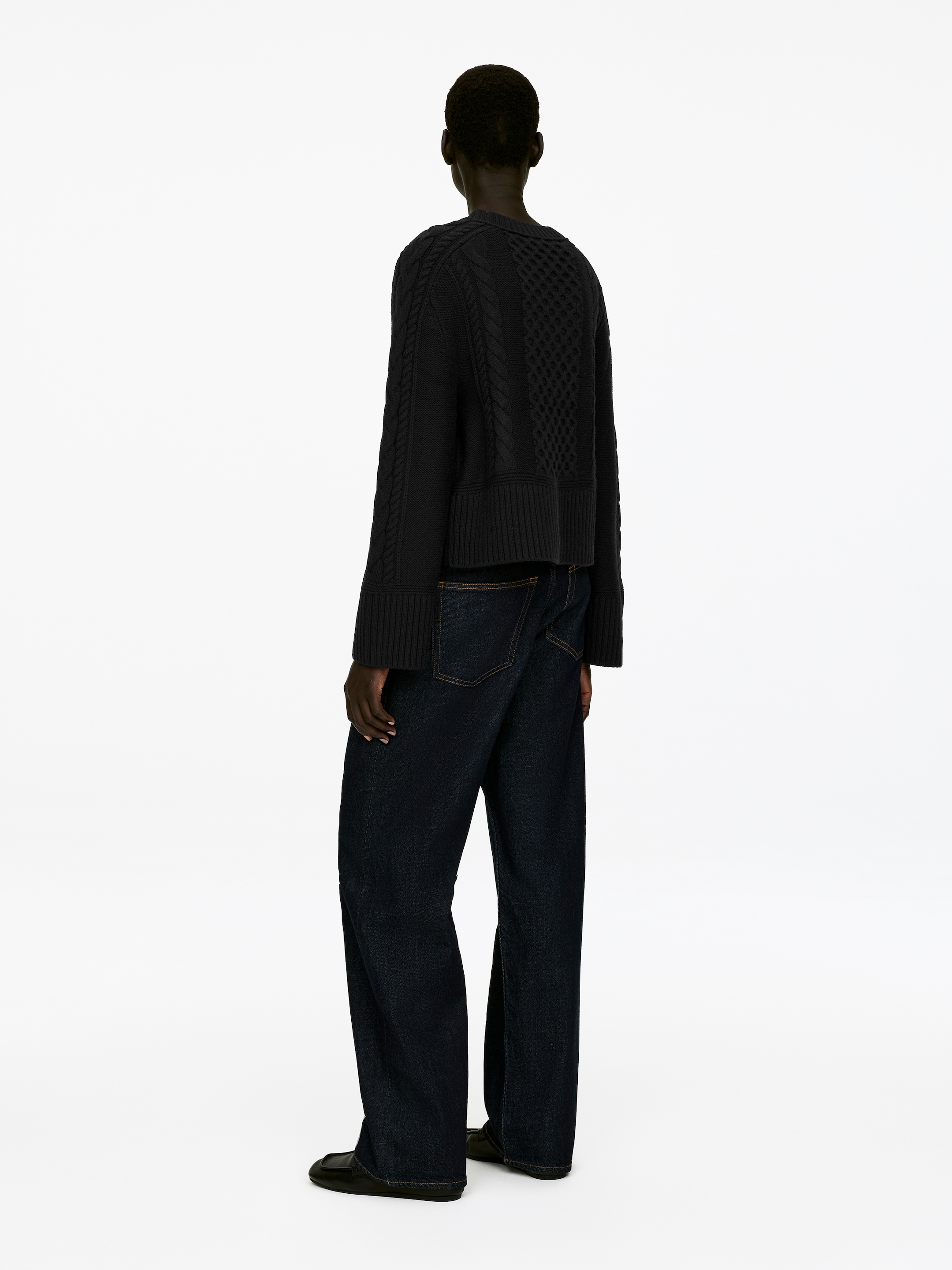 Grösseres Bild ansehen: Eine dunkle Strickjacke, mit Zopfmuster-Ärmeln, einem strukturierten Rückenpanel und gerippten Abschlüssen, wird vom Model getragen. Sie ist mit dunklen Jeans und schwarzen flachen Schuhen gestylt.