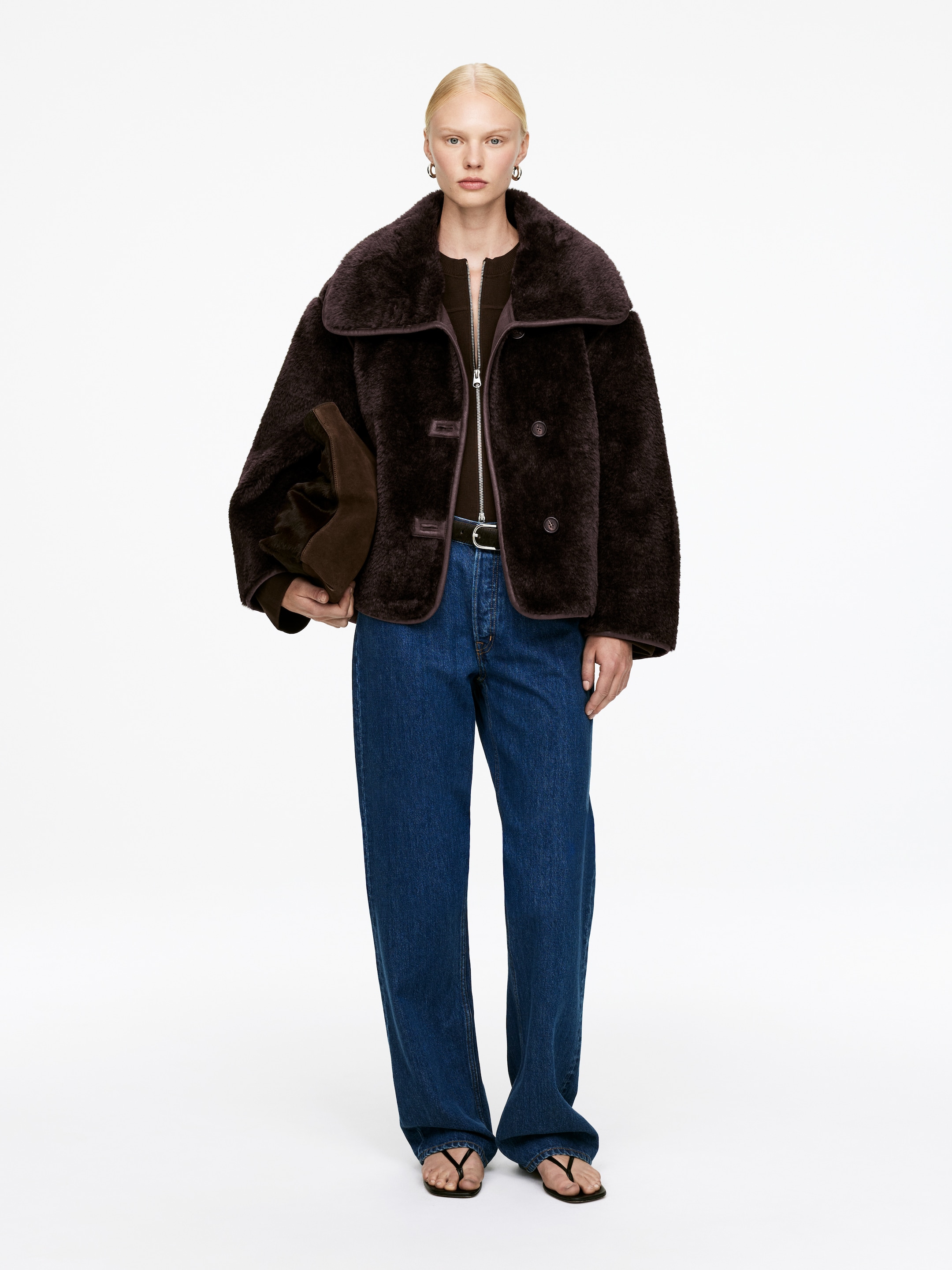 Zobrazit větší obrázek: Tmavě hnědá bunda ve stylu shearling je otočená dopředu, má široký, zaoblený límec a viditelný zip pod knoflíkovou légou. Představuje uvolněný, krabicový střih s kontrastním lemováním podél okrajů, vrstvený přes tmavý top a modré džíny.