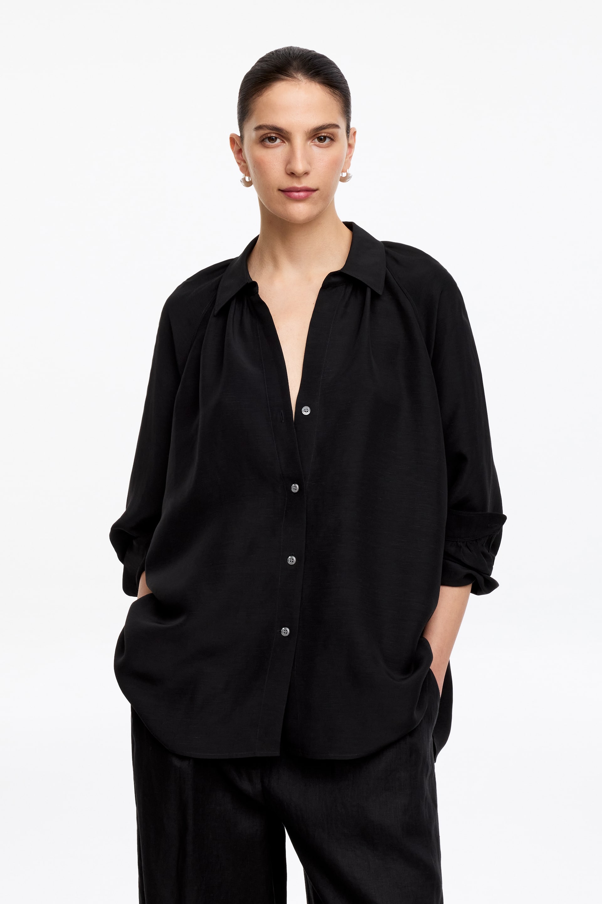 Ver imagen más grande: La modelo lleva puesta una camisa negra voluminosa con cuello con solapas, fruncidos y puños enrollados, dejando varios botones delanteros desabrochados. La modelo combina la camisa con pantalones oscuros.