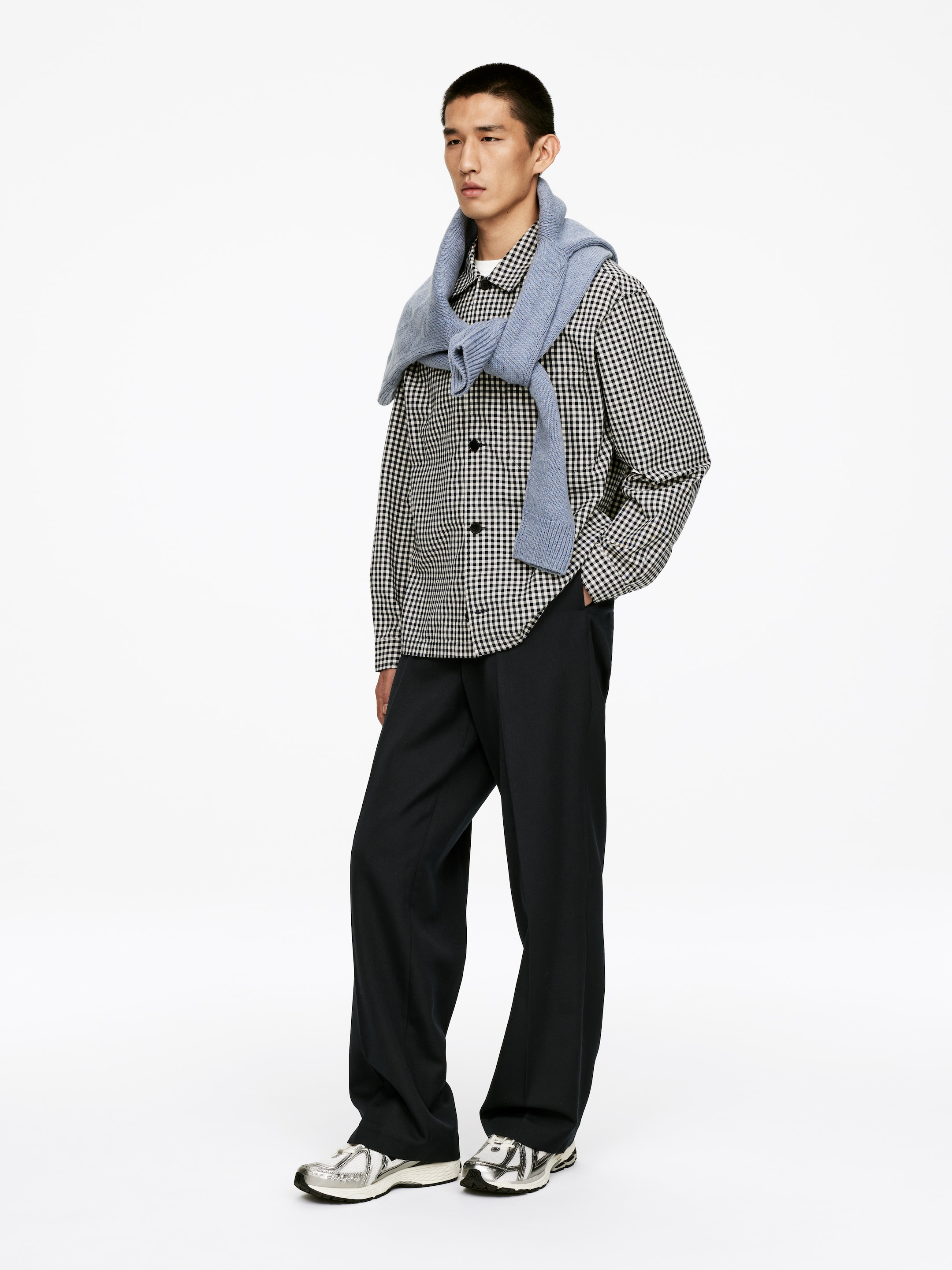 Visualizza immagine più grande: Una camicia a maniche lunghe in gingham bianco e nero viene indossata con pantaloni scuri a gamba larga e scarpe sportive argento. Un maglione a trecce azzurro chiaro è drappeggiato sulle spalle. L'insieme viene mostrato da un leggero angolo, evidenziando la vestibilità rilassata e il drappeggio dei capi.