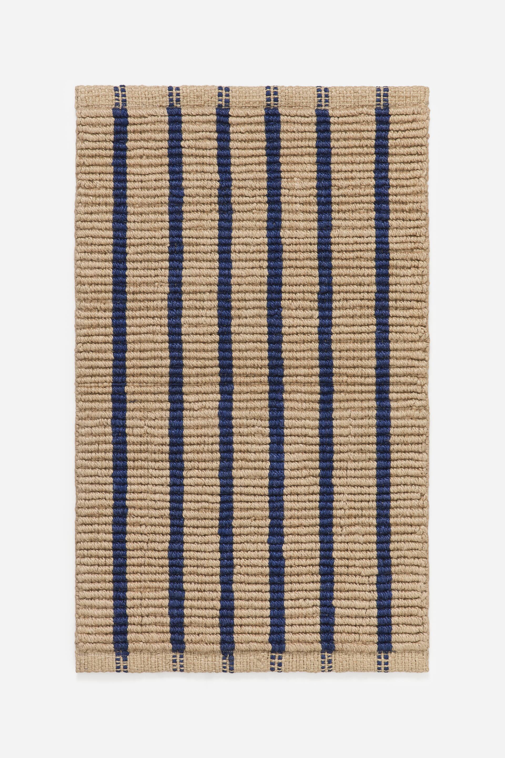 Visa större bild: En rektangulär matta visar en tätt vävd, naturligt ljusbrun textur. Vertikala marinblå ränder löper över mattan, med ändarna av varje rand som framträder som små, segmenterade block vid över- och underkanten.
