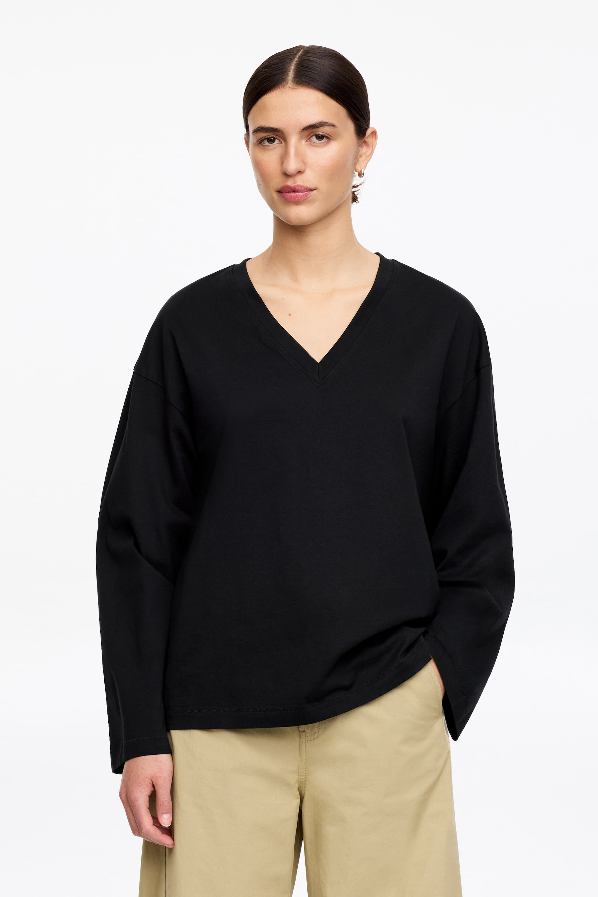 Agrandir l'image: Un haut noir à manches longues, coupe ample, avec un col en V et un ourlet droit est présenté, assorti à un pantalon kaki et une main reposant dans une poche pour montrer son drapé à hauteur des hanches.