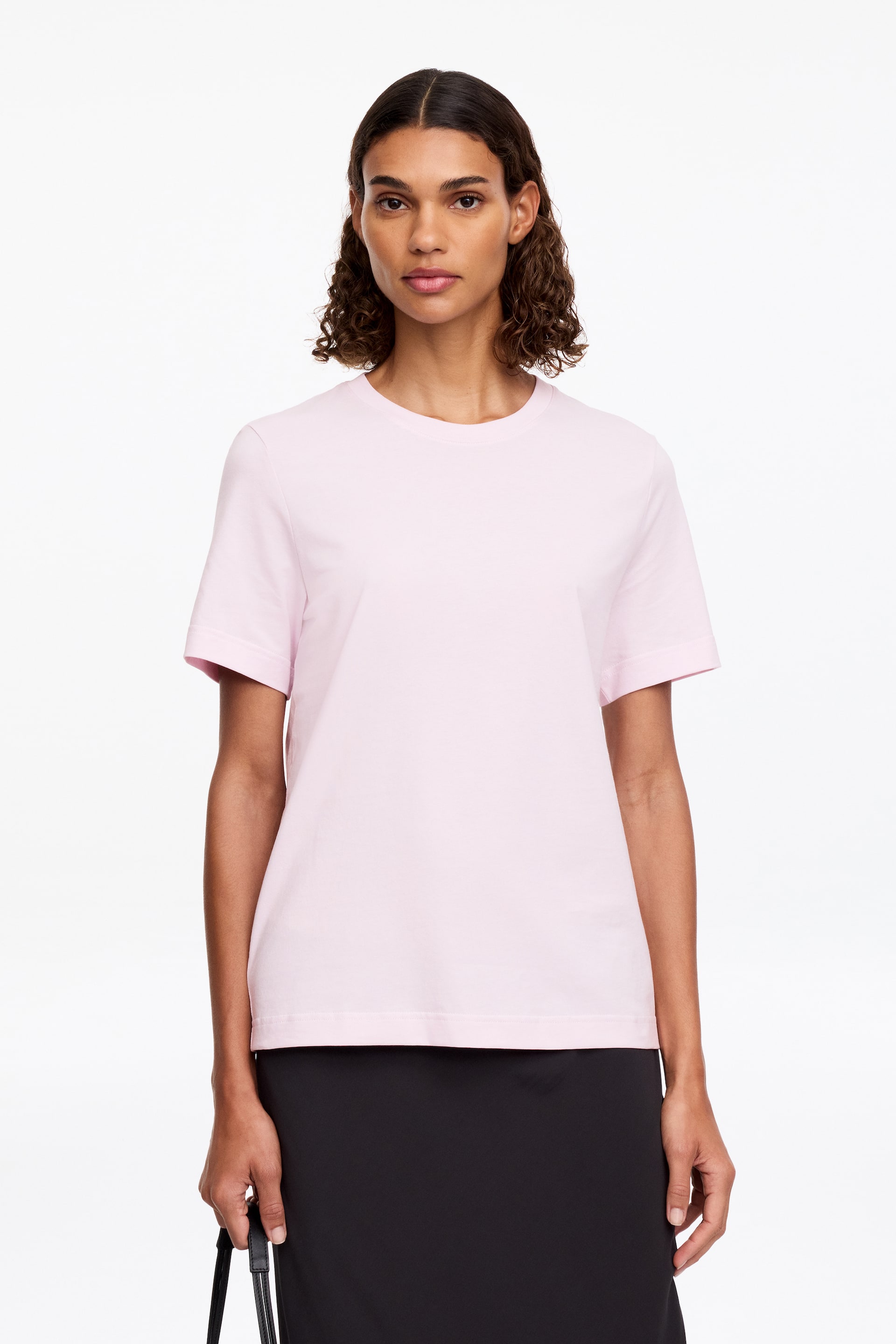 Grösseres Bild ansehen: Das Model trägt ein hellrosa T-Shirt. Es hat einen gerippten Rundhalsausschnitt und kurze Ärmel und wird mit einem schwarzen Rock kombiniert.