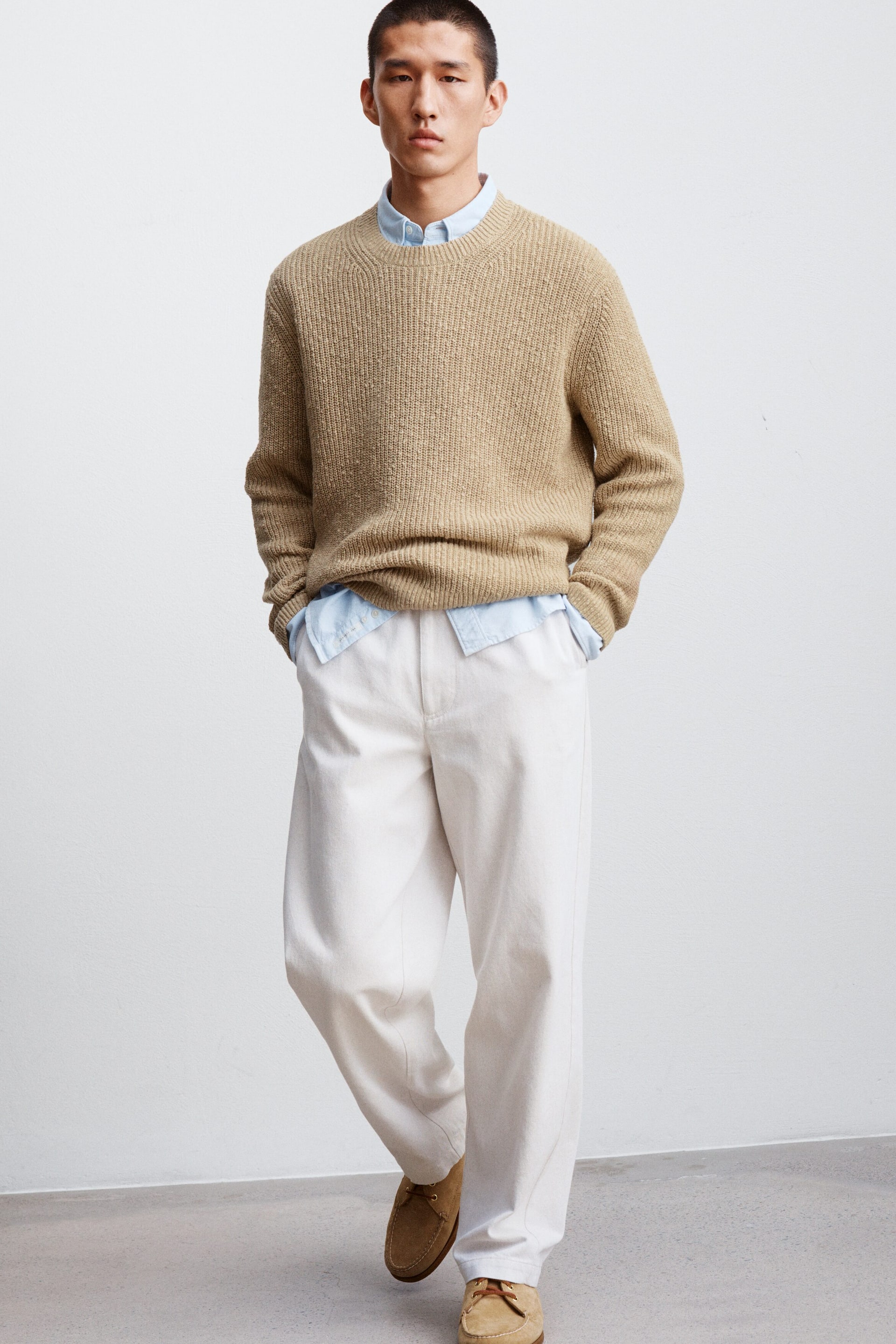 Se større billede: Den beige chunky strikkede crew neck sweater har en fremtrædende ribbet struktur overalt, med lange ærmer, ribbede manchetter og en ribbet kant. En lyseblå skjorte med krave er synlig under sweateren.