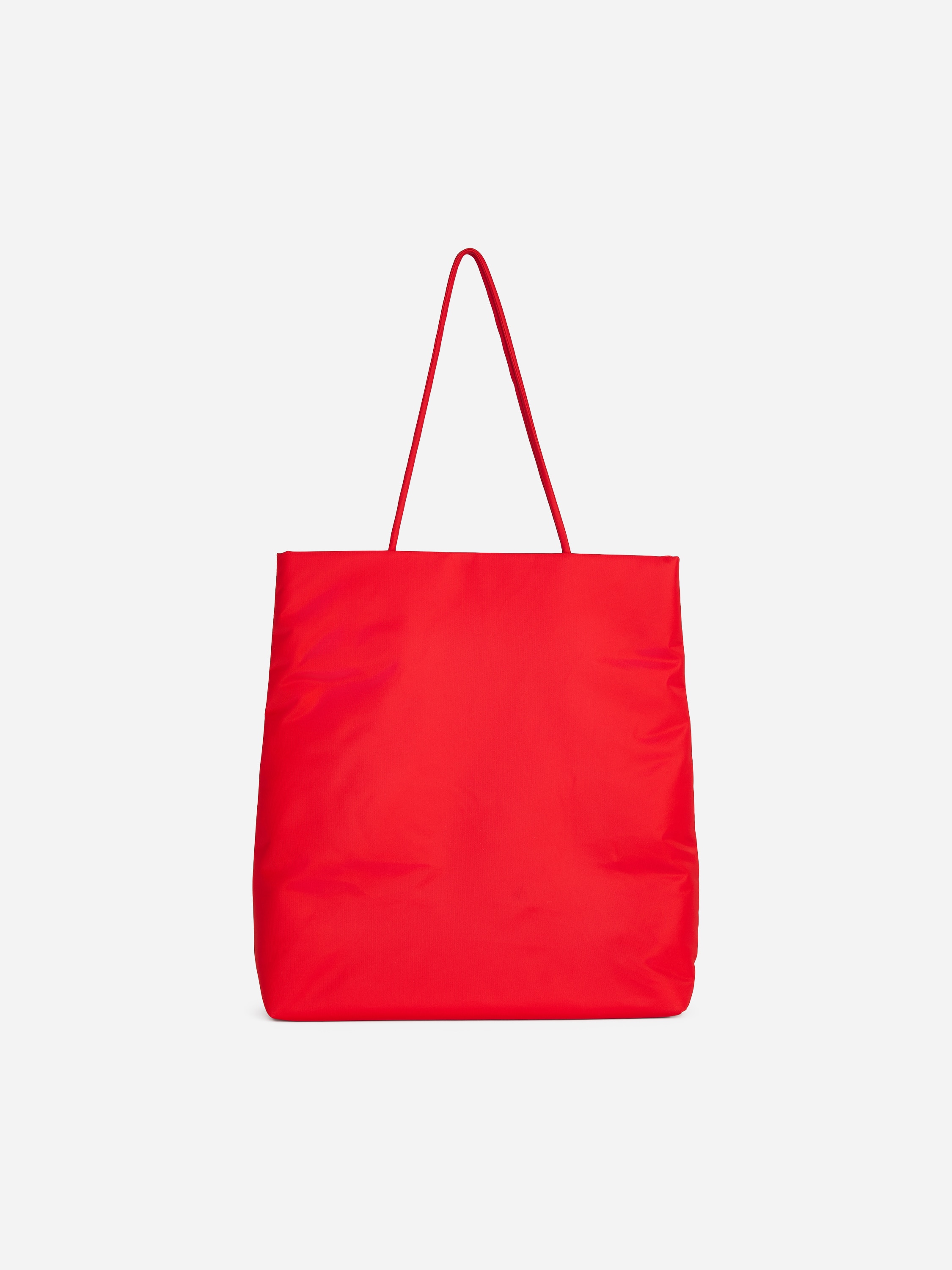A bright red, padded nylon tote bag with thin, rounded shoulder straps is being carried over the modelâs shoulder. The model is wearing a light beige trench coat, with the rectangular bag positioned under the arm.