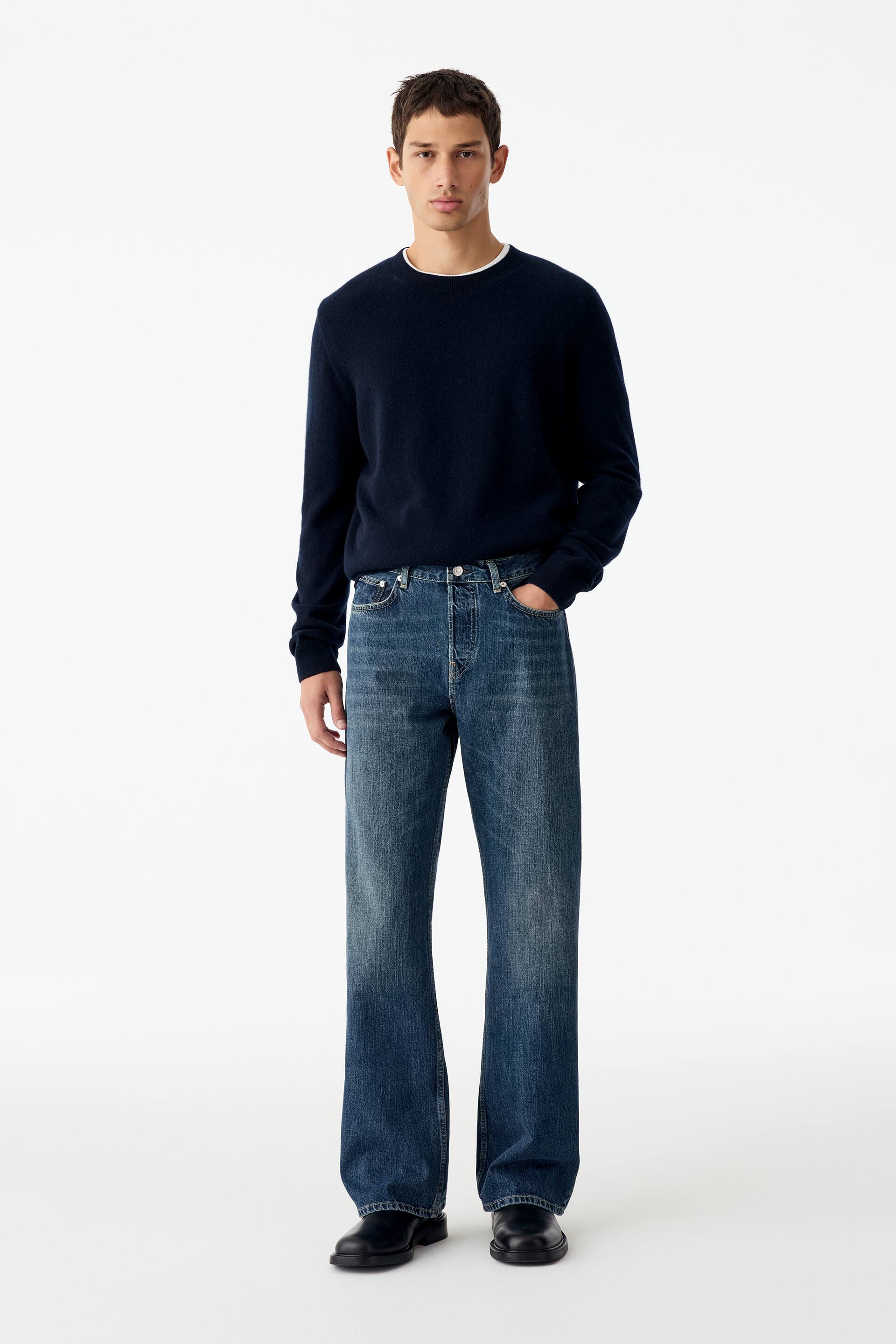 Agrandir l'image: Un jean en denim bleu à taille mi-haute, coupe regular et jambes bootcut est porté par le mannequin, qui se tient debout avec une main posée dans la poche avant. Le jean est associé à un pull à col rond foncé et des bottes en cuir noir.