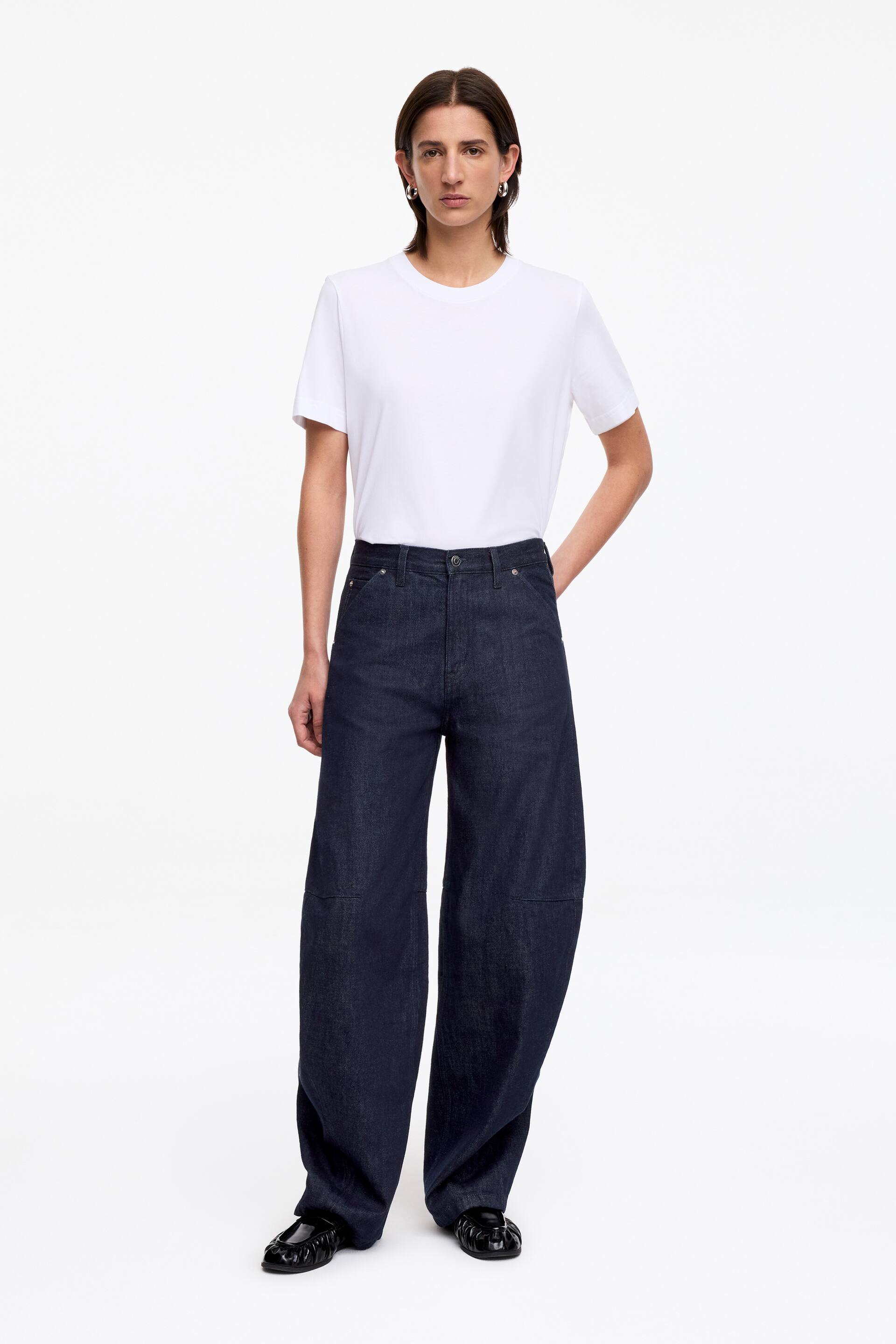 Προβολή μεγαλύτερης εικόνας: Σκούρο πλύσιμο, ψηλόμεσο, τζιν παντελόνι με φαρδιά μπατζάκια παρουσιάζεται από μπροστά. Το τζιν διαθέτει σιλουέτα με μπατζάκια σε σχήμα βαρελιού με ραφές στις γόνατες και συνδυάζεται με λευκό μπλουζάκι με στρογγυλή λαιμόκοψη.