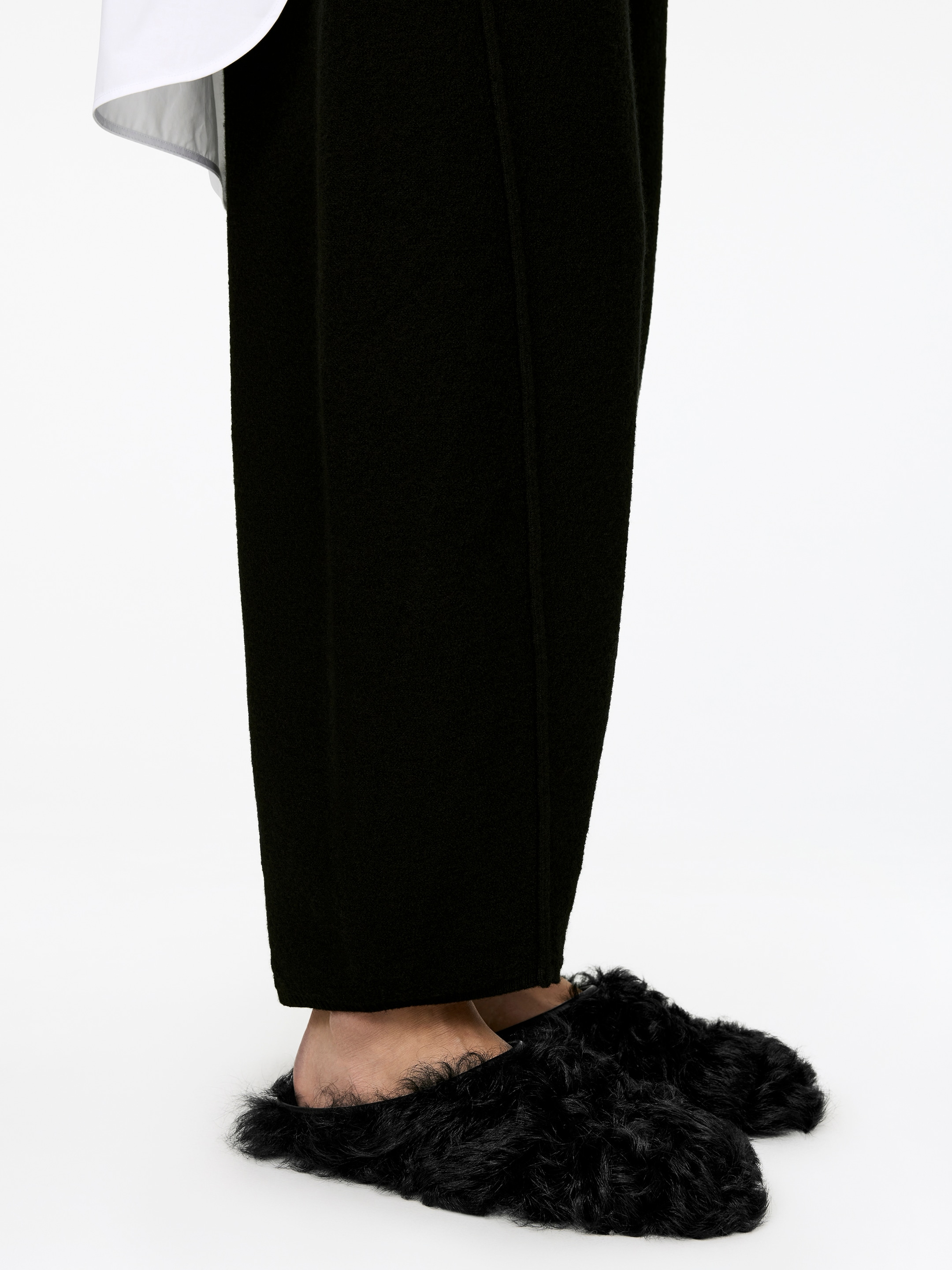 Agrandir l'image: Les mules en shearling noir et doux sont portées par le mannequin. Les mules présentent une texture ébouriffée et un design à l'arrière ouvert, et le pantalon noir du mannequin repose sur leur partie supérieure.