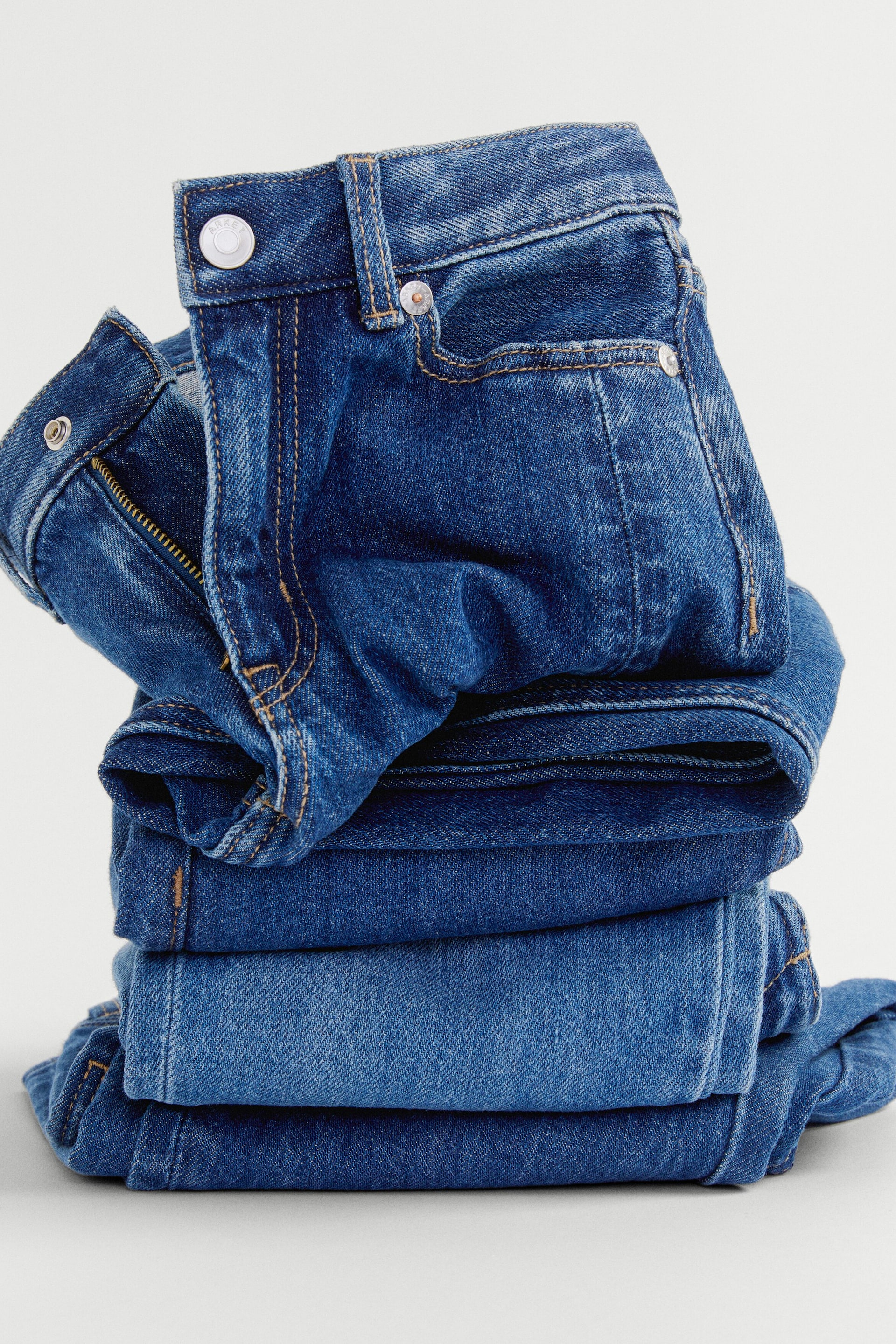 Visa större bild: Ett staplat set med vikta blå denimjeans visas upp, där det översta paret är vänd framåt och visar knäppning med knapp, dragkedjegylf, bälteshällor samt framfickor med metalnitar. Denimen uppvisar varierande nyanser av blått.