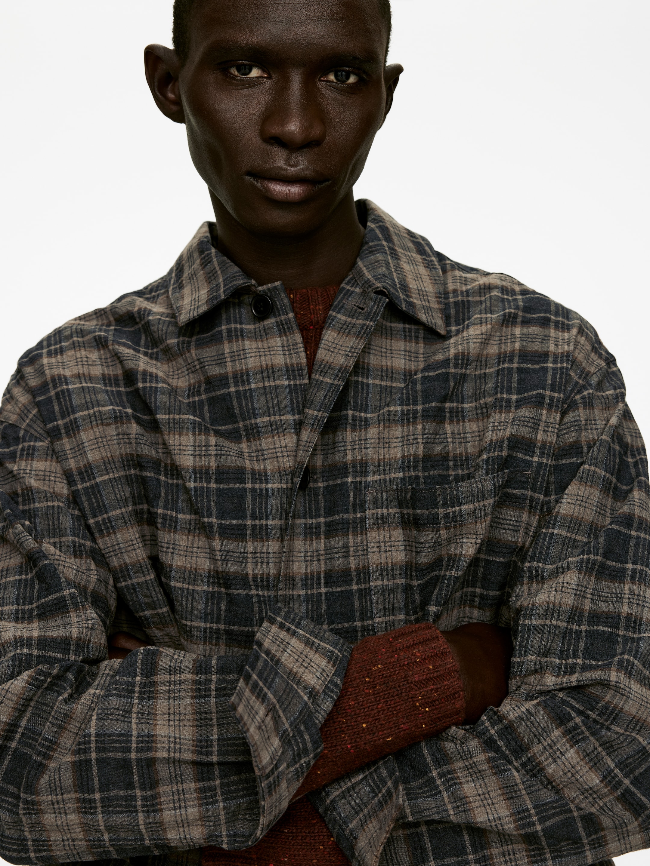 Grotere afbeelding bekijken: Een geruite overshirt, met een grijs, bruin en donkerblauw ruitpatroon, wordt gedragen vanuit een vooraanzicht, iets schuin. Het heeft een puntige kraag en een enkele zichtbare knoopsluiting aan de bovenkant. Een roodbruin, gespikkeld trui met een geribbelde textuur wordt eronder gedragen, waarbij de mouwen zichtbaar zijn bij de onderarmen.