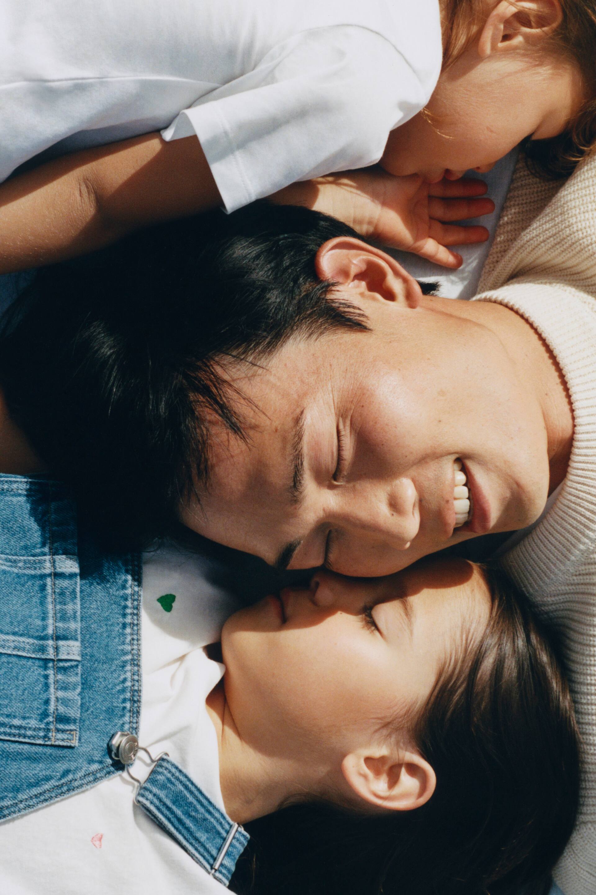 Se større billede: En cremefarvet striktrøje viser en ribbet struktur og en rund halskrave. En hvid t-shirt med et lille grønt hjertebroderi på brystet er lagdelt under blå denimoveralls med metalspænder.