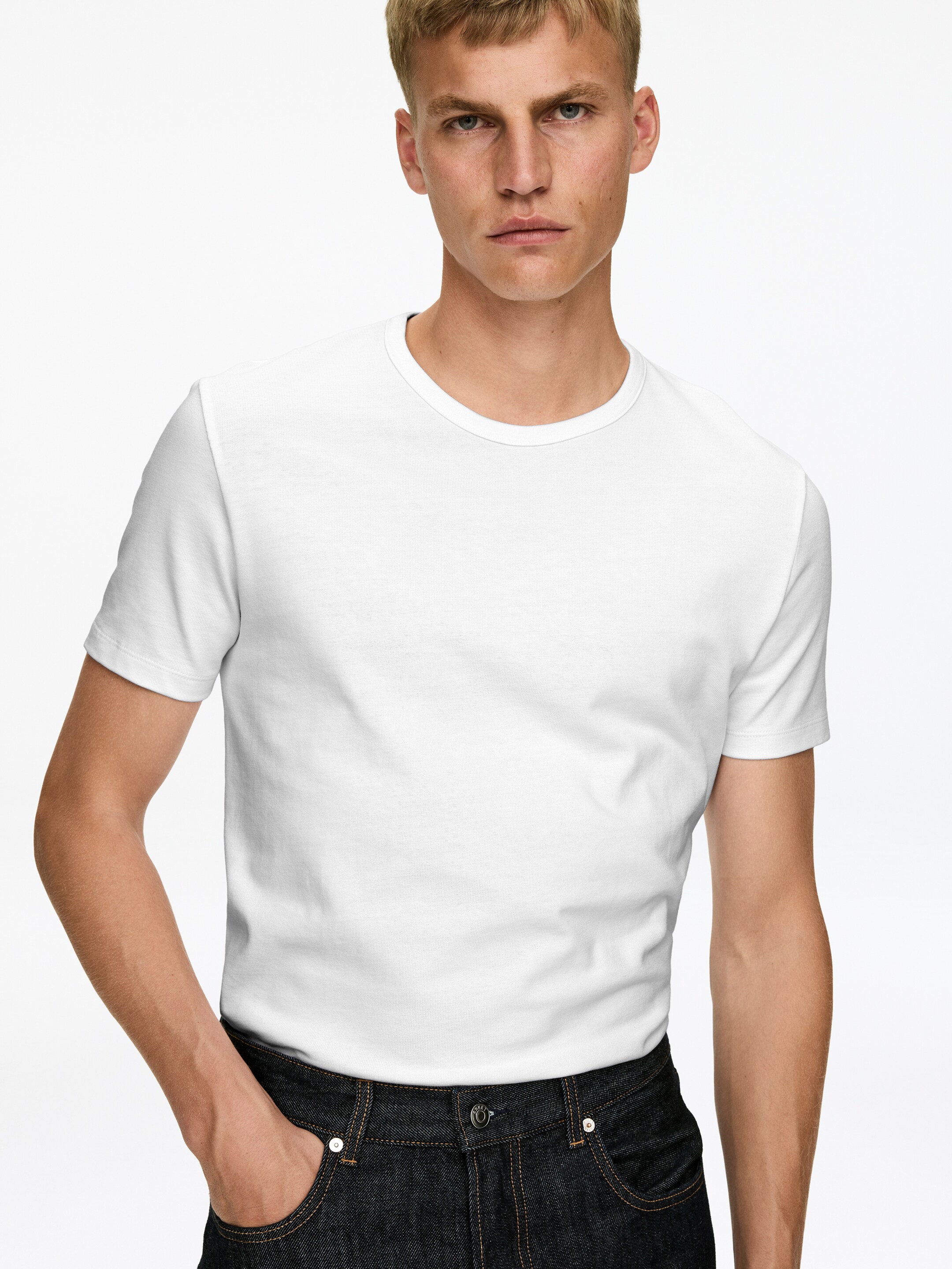 Agrandir l'image: Un t-shirt blanc à col rond est présenté face avant. Il se caractérise par des manches courtes et une silhouette ajustée, et est stylé en étant rentré dans un jean foncé.