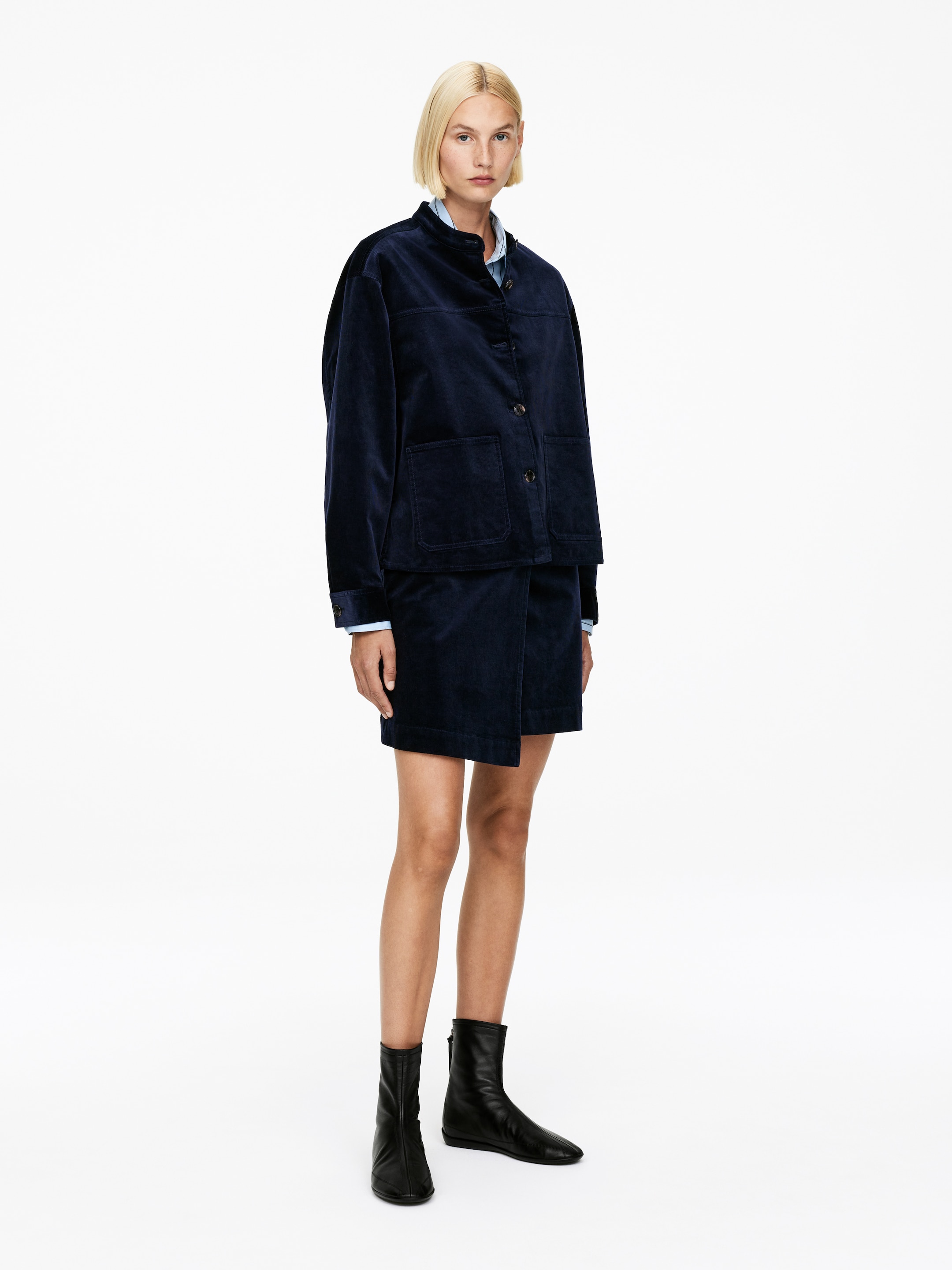 Ver imagen más grande: Una chaqueta de pana azul oscuro y una falda asimétrica de estilo envolvente a juego están siendo modeladas. La chaqueta cuadrada presenta un cuello de banda, cierre frontal con botones oscuros y dos grandes bolsillos de parche, con una camisa de cuello azul claro en capas debajo, visible en el cuello y los puños. Unas botas de cuero negro hasta el tobillo con suela plana y punta puntiaguda completan el conjunto.