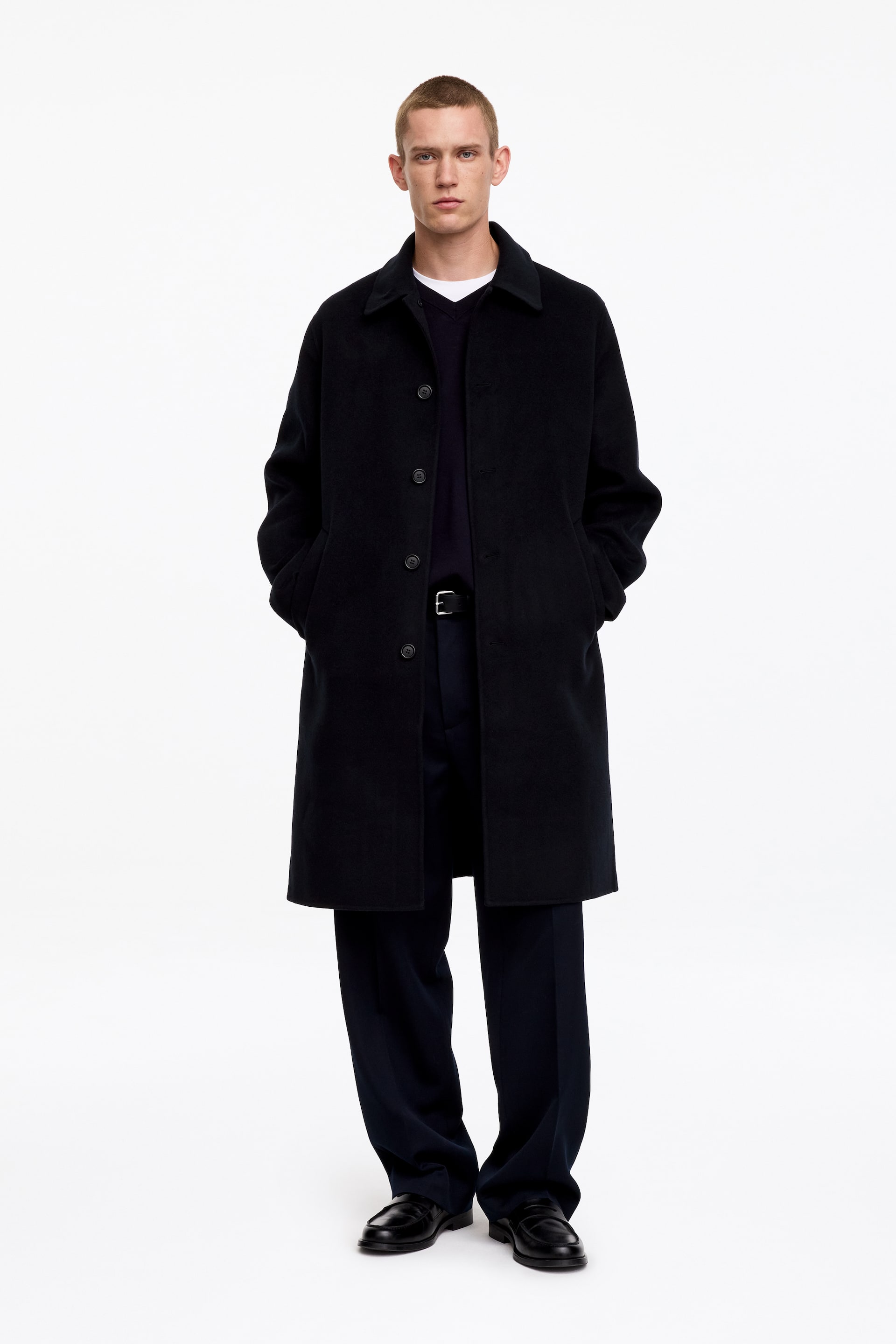 View larger image: A dark navy wool coat, cut for a relaxed fit, is being worn by the model. The coat features a regular collar, front button fastening, and slanted front pockets where the model has her hands placed.