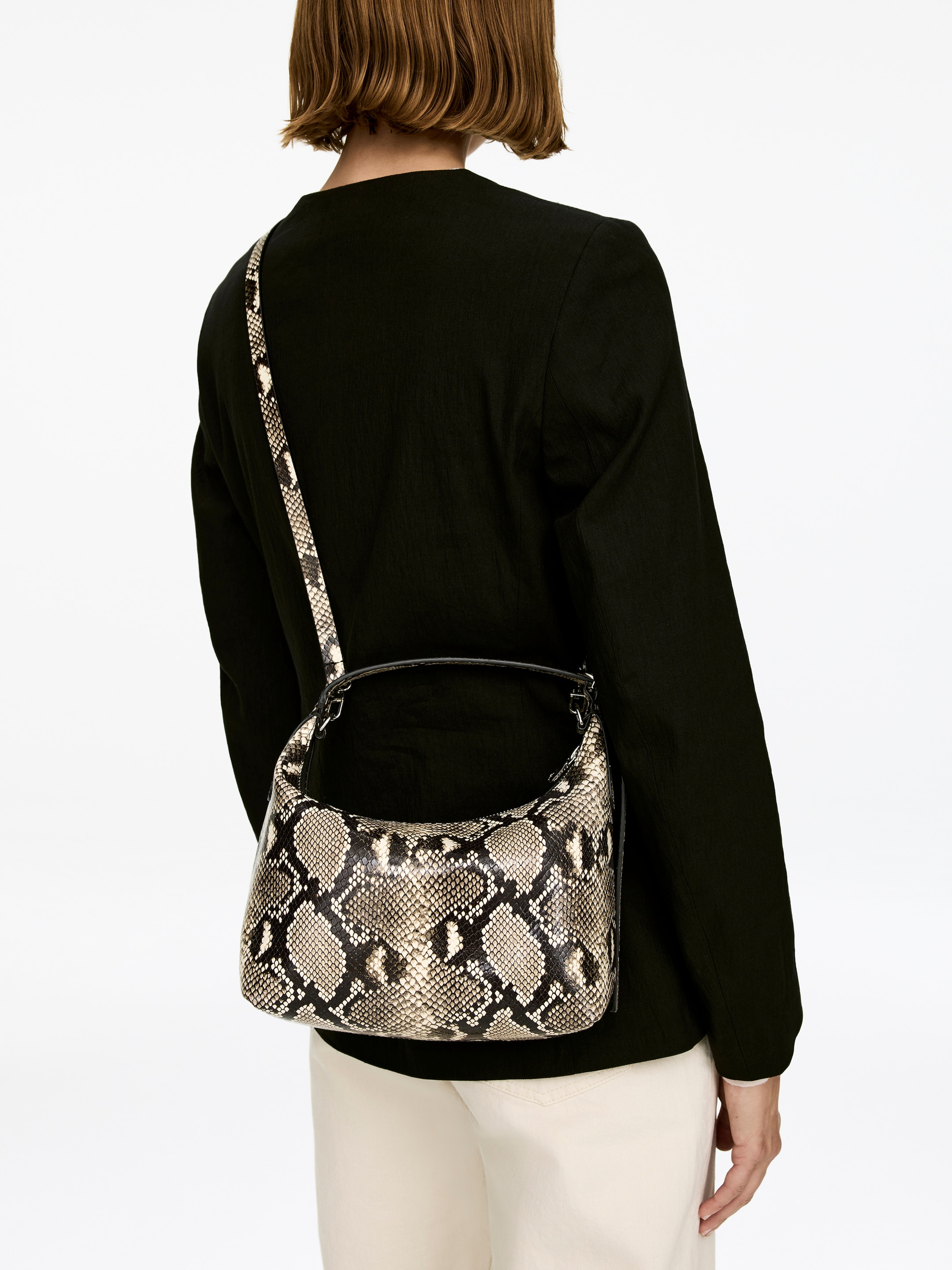 Ver imagen más grande: Un bolso de cuero cuadrado con un patrón de piel de serpiente está siendo llevado por la modelo. El bolso presenta un asa superior y una correa de hombro delgada y desmontable que cruza la espalda de la modelo sobre una chaqueta negra.
