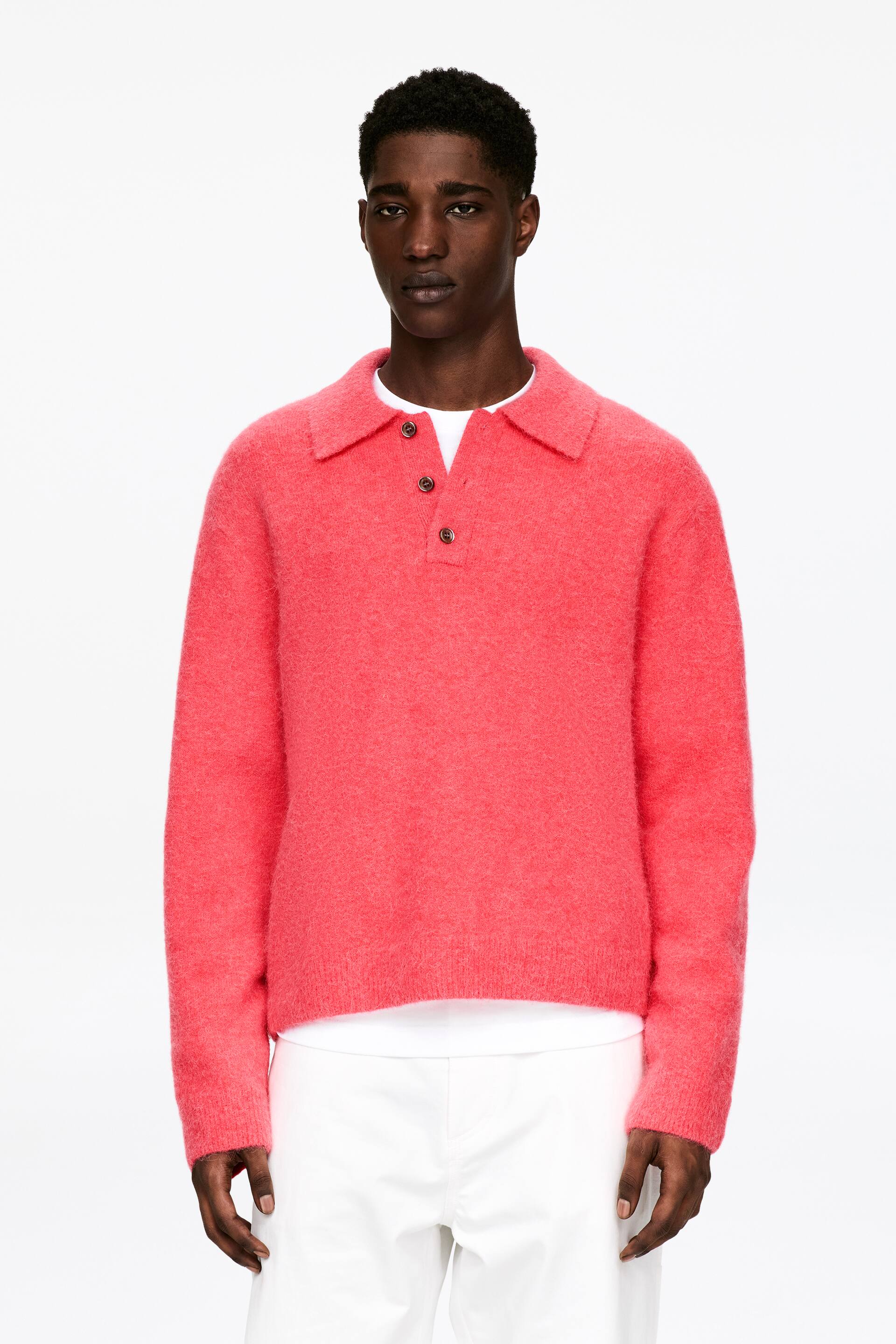 Zobacz większy obraz: Modelka ma na sobie koralowy, gładko dziergany polo z włochatą powierzchnią, nałożony na białą koszulkę. Ta luźno dopasowana koszulka posiada kołnierzyk, półplisę z trzema guzikami w kolorze brązowym oraz ściągacze przy mankietach i na dole.