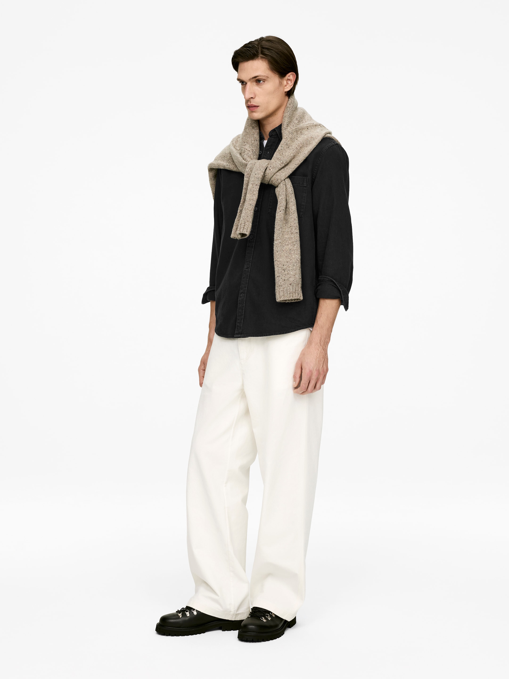 Agrandir l'image: Une chemise noire à manches longues et boutonnée est portée, légèrement tournée vers la droite. Ses manches sont retroussées jusqu'aux avant-bras et un pull en tricot beige clair est drapé sur ses épaules.