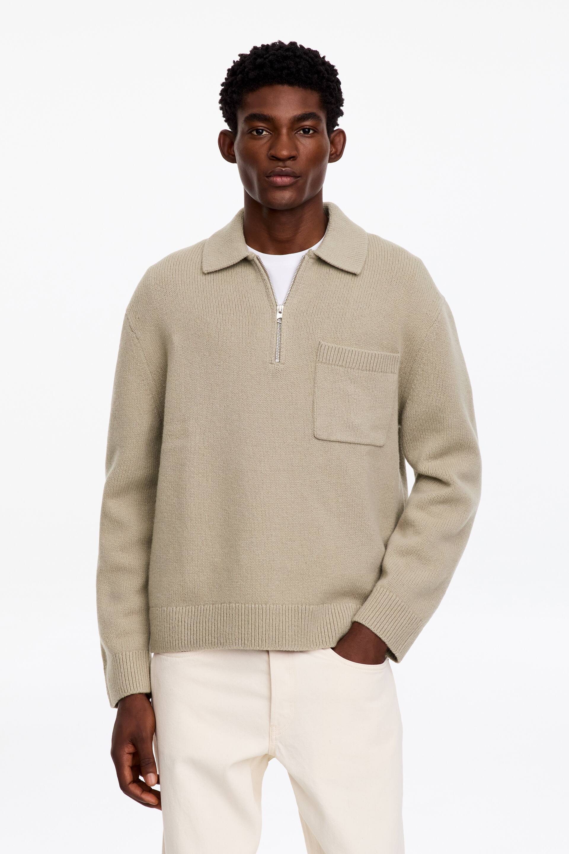 View larger image: A light beige knit jumper featuring a half-zip collar, a chest patch pocket, and ribbed cuffs and hem is being worn by the model. The jumper is layered over a white crew-neck T-shirt and styled with off-white trousers.