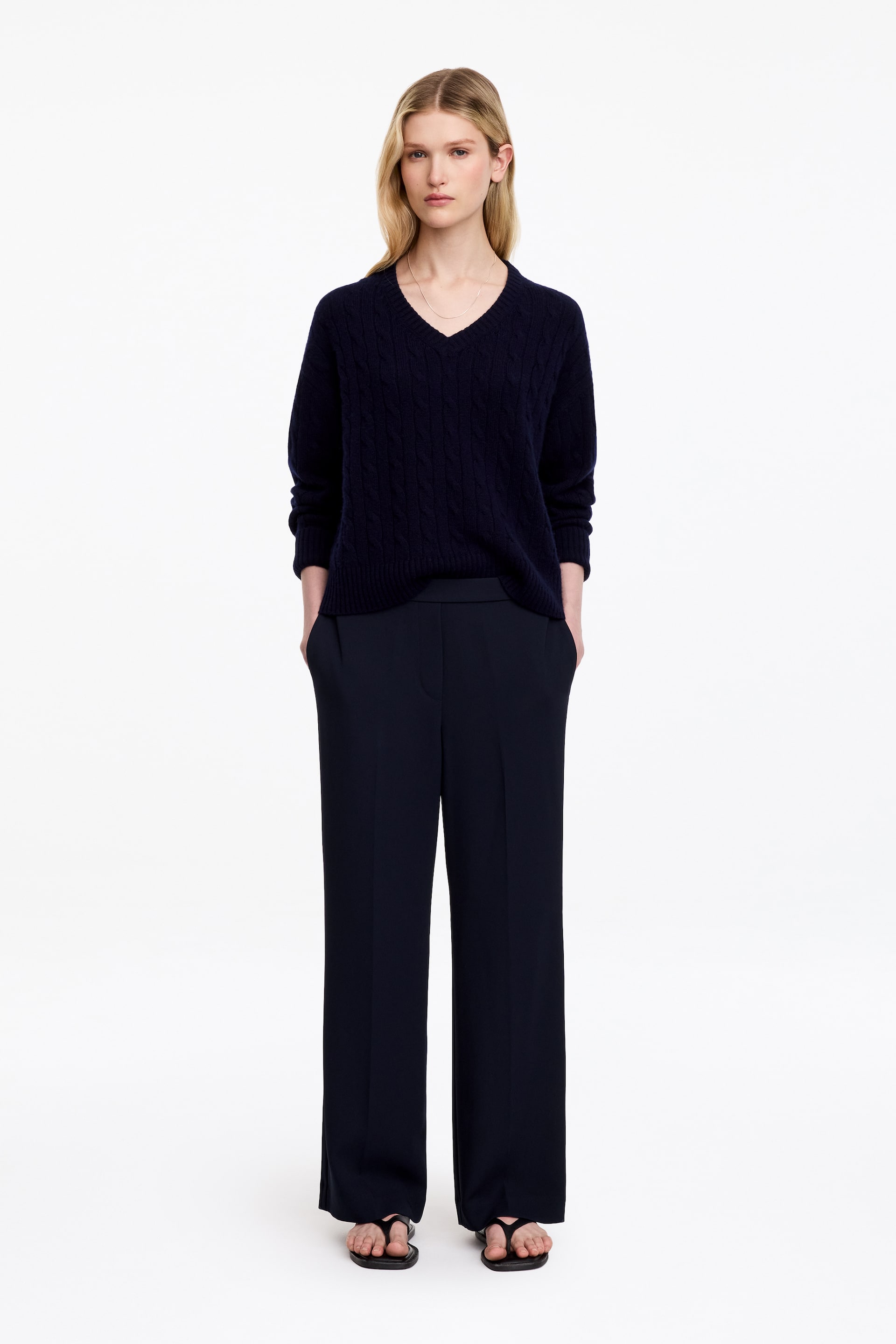 Visualizza immagine più grande: Pantaloni blu navy scuro caratterizzati da una silhouette a gamba dritta, pieghe anteriori stirate e tasche laterali indossati dalla modella. La modella è in piedi con le mani inserite nelle tasche, abbinando i pantaloni a un maglione scuro con scollo a V coordinato e sandali bassi.