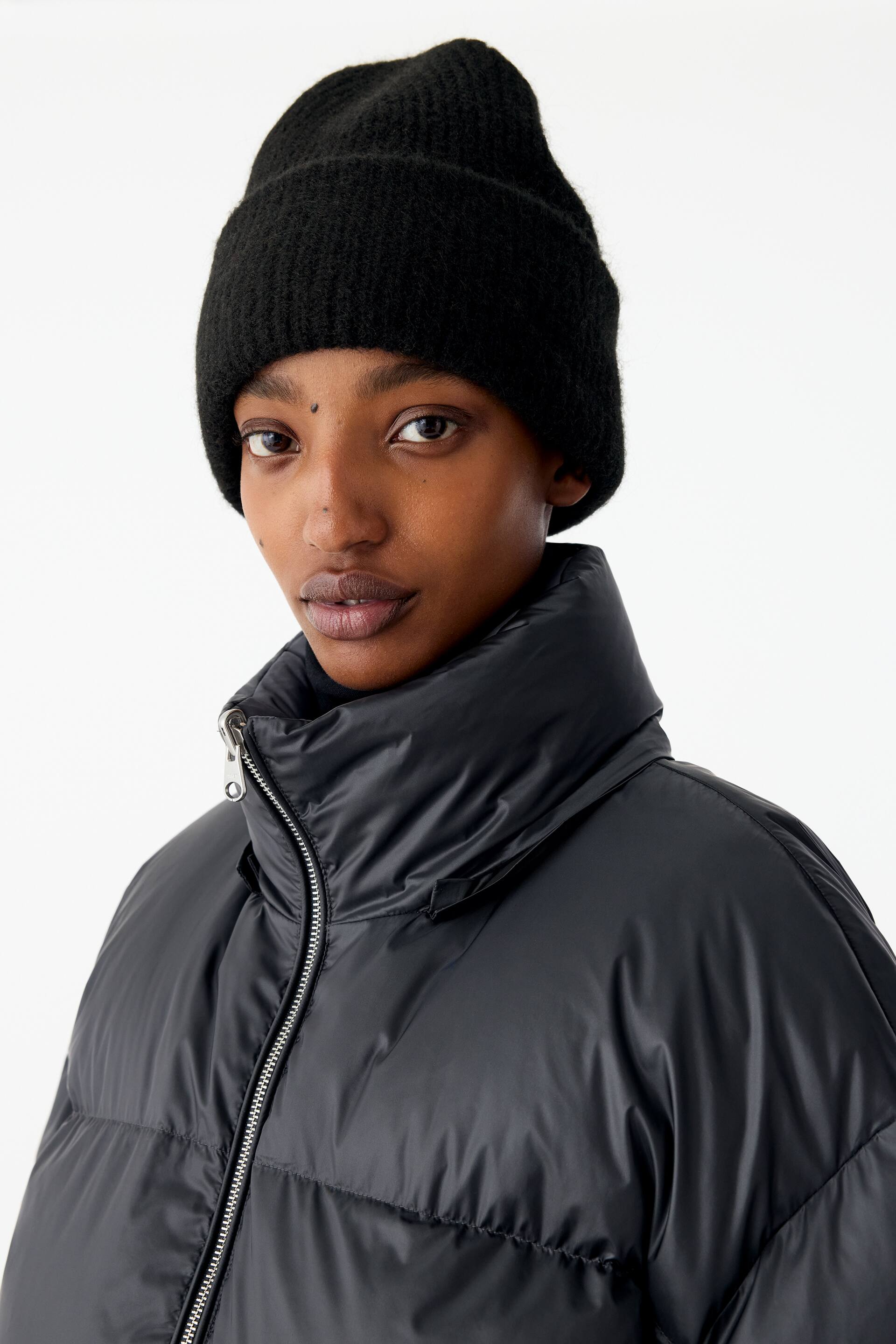 Grotere afbeelding bekijken: Een zwarte pufferjas met een hoge opstaande kraag en een zilverkleurige ritssluiting aan de voorkant wordt gedragen door het model. Ook draagt het model een zwarte gebreide beanie op het hoofd.