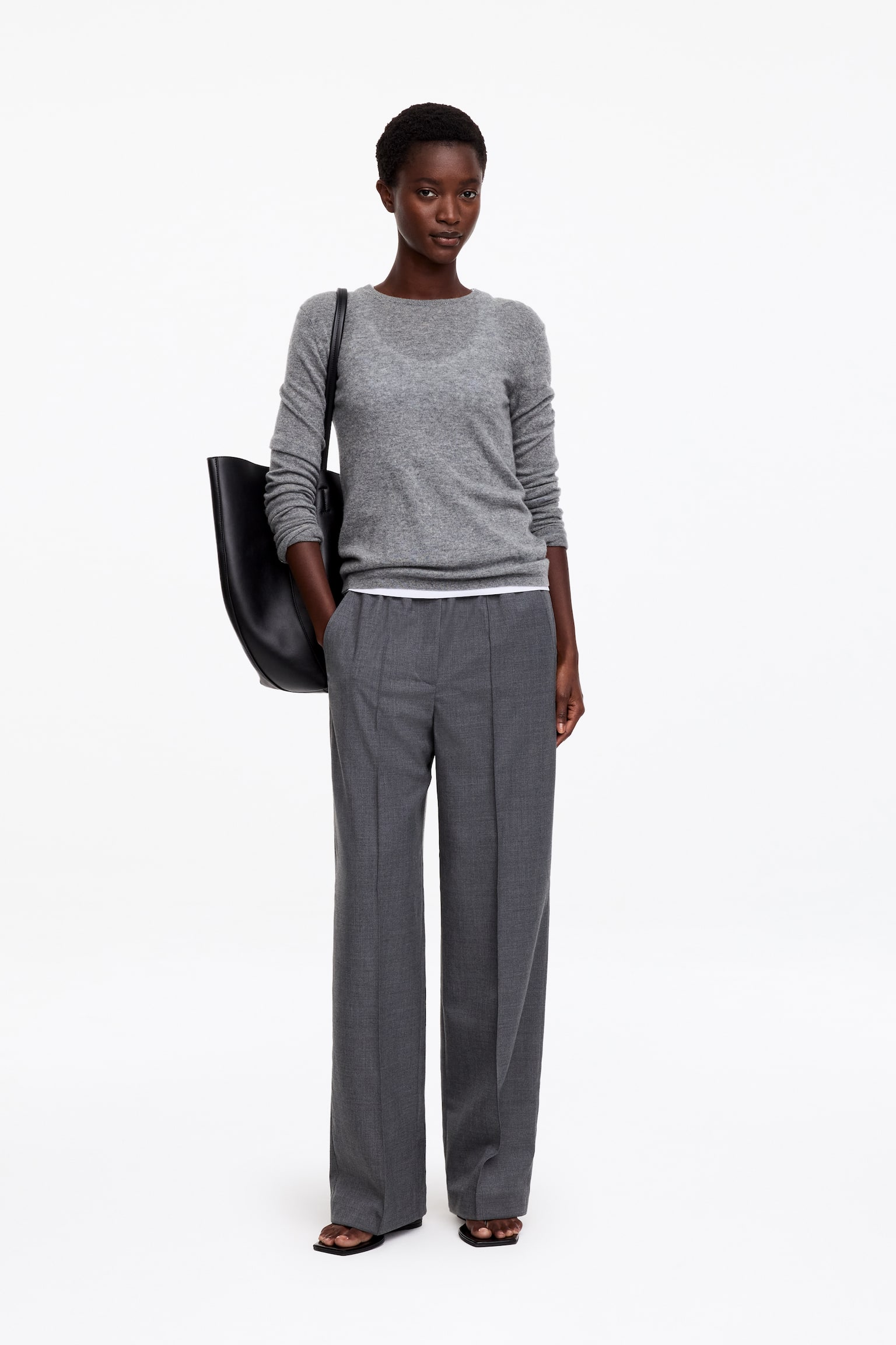 Grey straight-leg trousers with an elasticated waist and sharp front creases are worn by the model, who is standing with one hand resting in a side pocket. The trousers are styled with a grey crew-neck sweater, a white undershirt visible at the hem, and a black shoulder bag. - 1