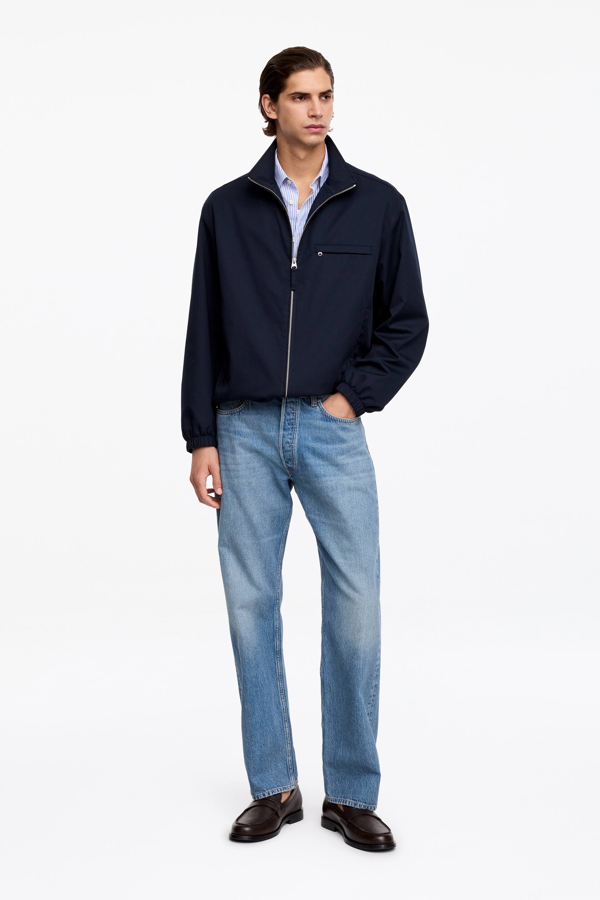 Agrandir l'image: Un blouson bleu marine est présenté de face, doté d'un col montant, d'une fermeture éclair centrale de couleur argentée, d'une poche zippée horizontale sur la poitrine gauche, ainsi que de poignets et d'un ourlet élastiqués. Il est porté par-dessus une chemise à col rayée bleu clair et blanche.