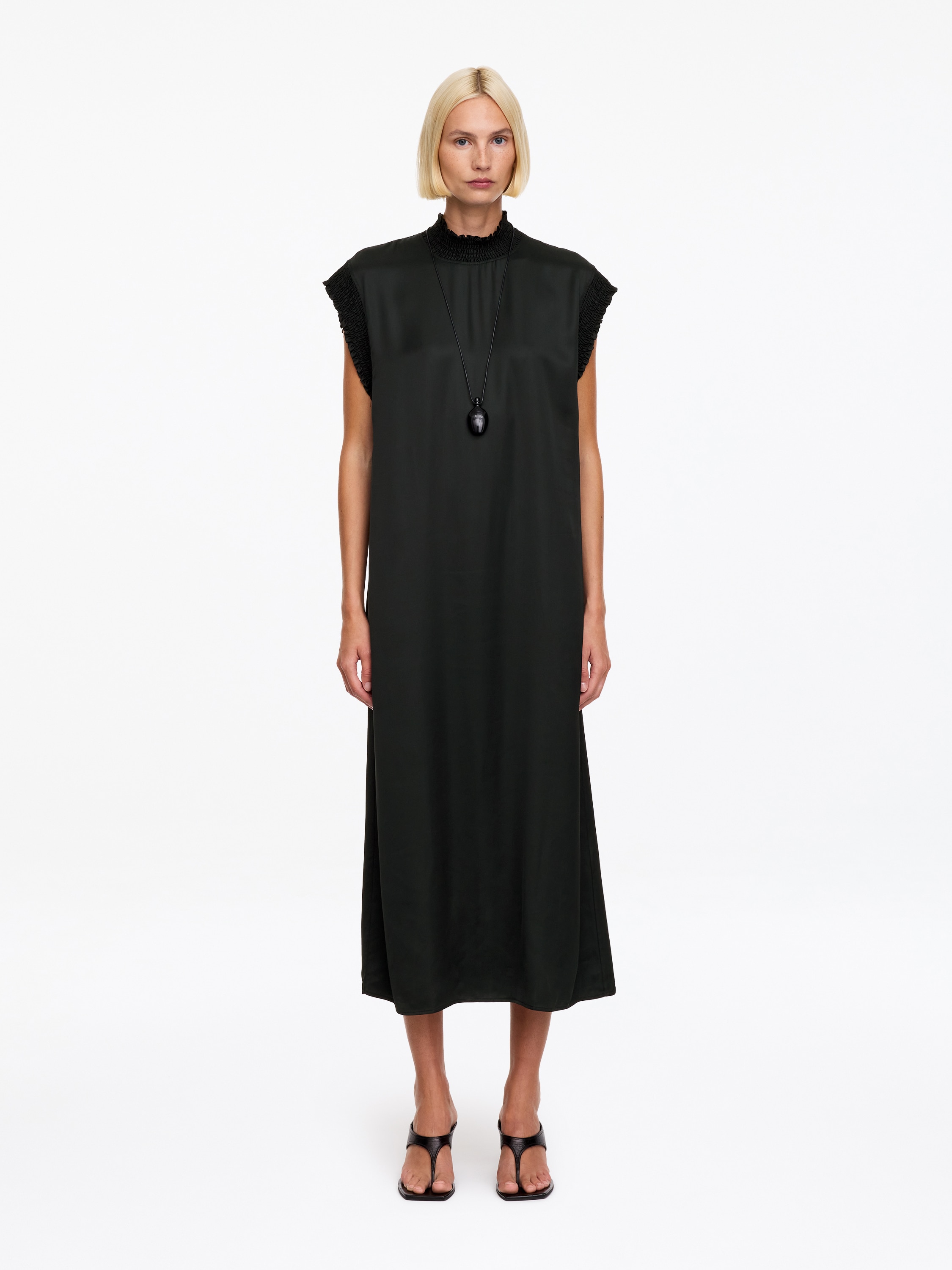 Um vestido preto sem mangas Ã© mostrado de frente, apresentando uma silhueta reta e descontraÃ­da com uma gola alta tipo mock neck. O vestido foi concebido com pormenores texturizados em smocking Ã  volta da gola e das cavas.