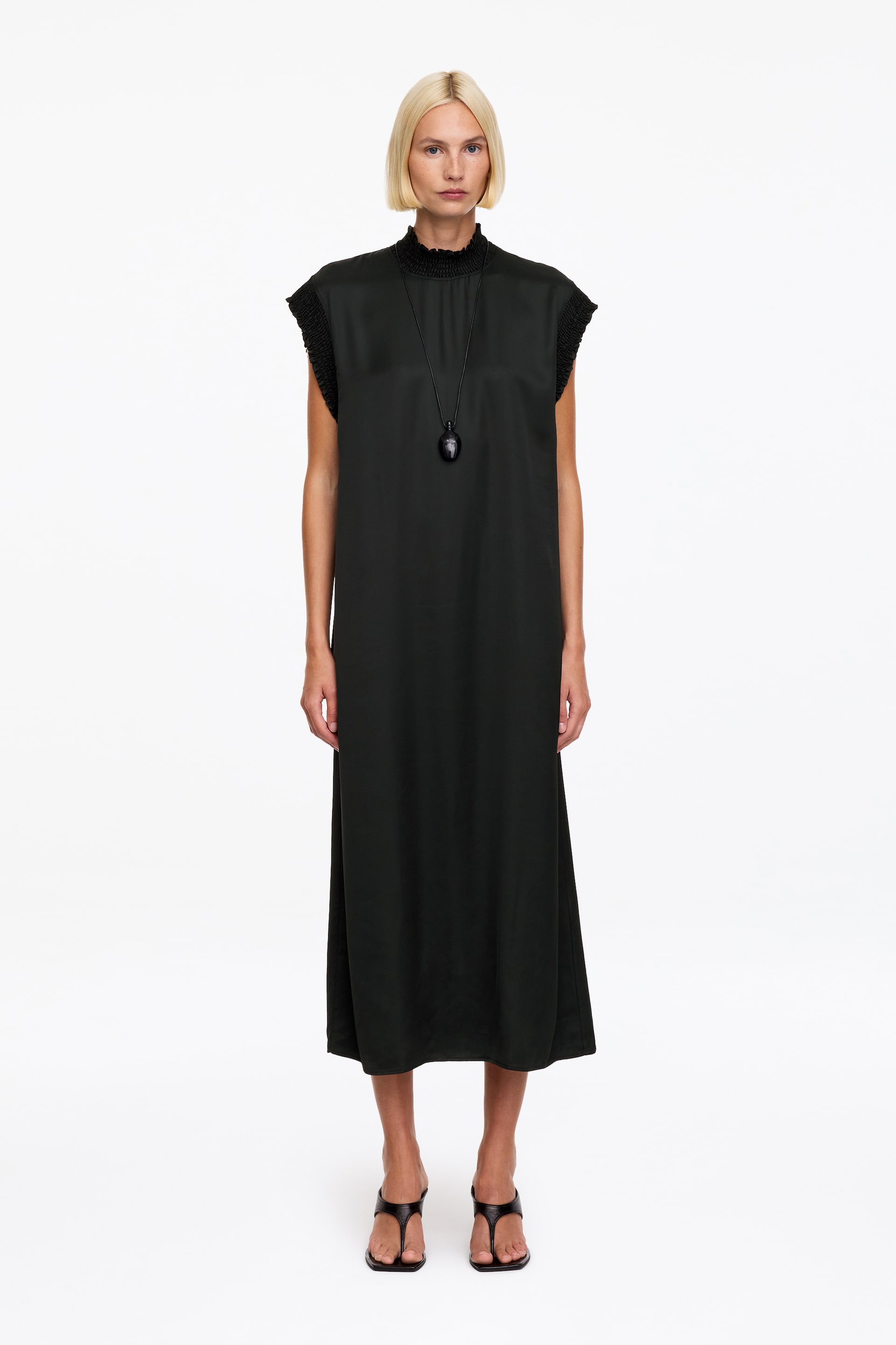Ver imagen más grande: La modelo luce un vestido midi negro sin mangas de corte relajado, con detalle de fruncido elástico en el cuello alto y en los bordes de las sisas. El vestido se combina con un collar largo negro con colgante y sandalias negras tipo tanga.