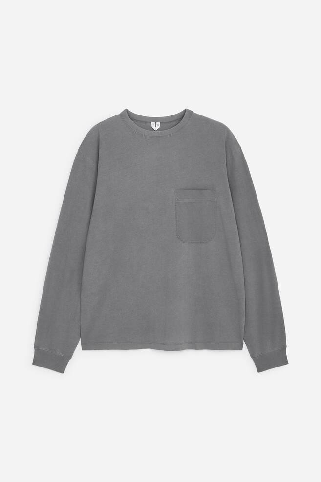 Ein graues Langarm-Sweatshirt mit Rundhalsausschnitt wird von vorne gezeigt. Es zeichnet sich durch eine lockere Passform, eine aufgesetzte Brusttasche auf der linken Seite, gerippte Bündchen und einen geraden Saum aus. - 1