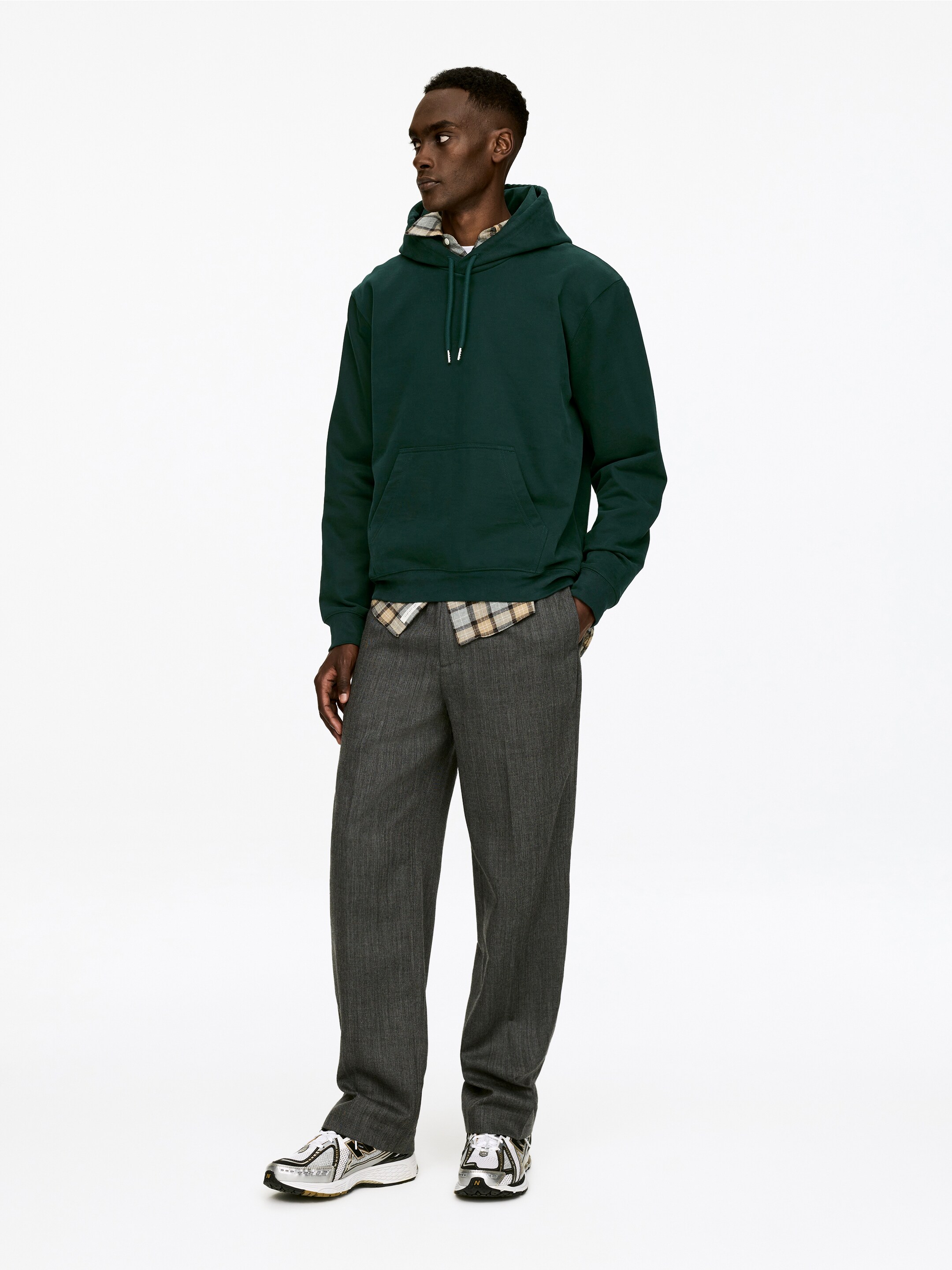 Ver imagem maior: Um hoodie verde escuro está a ser usado, ligeiramente voltado para a direita, e possui um capuz com cordão, um bolso canguru frontal e punhos e bainha canelados. Uma camisa aos quadrados está em camadas por baixo, com a gola e a bainha inferior à mostra.