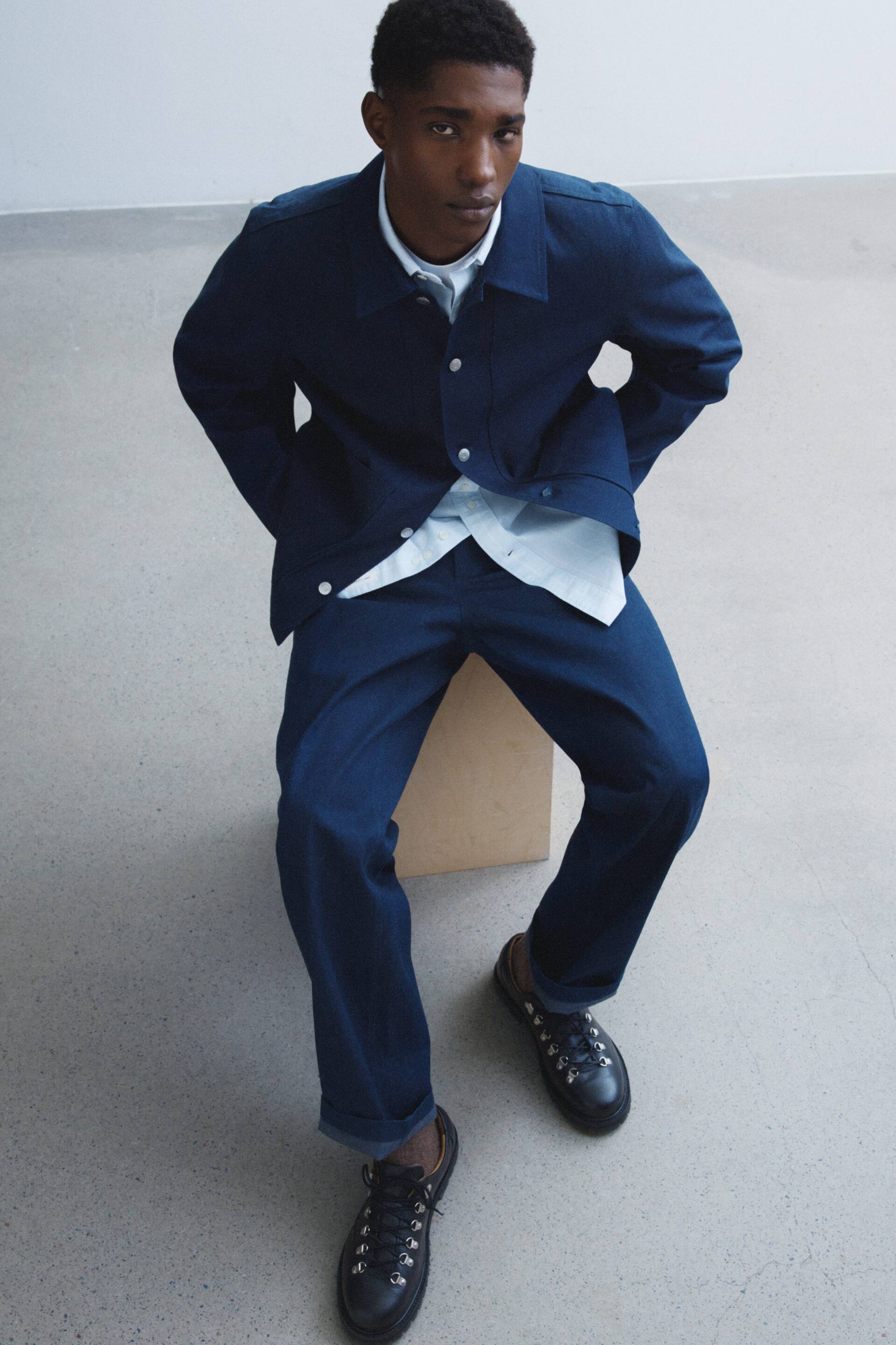 Vis større bilde: En marineblå jakke med spredt krage, knappelukking foran og lange ermer, bæres åpen over en lyseblå skjorte med krage. Matchende marineblå bukser komplementerer jakken, og presenterer en avslappet passform med oppbrettede kanter, mens de bæres av en sittende figur.