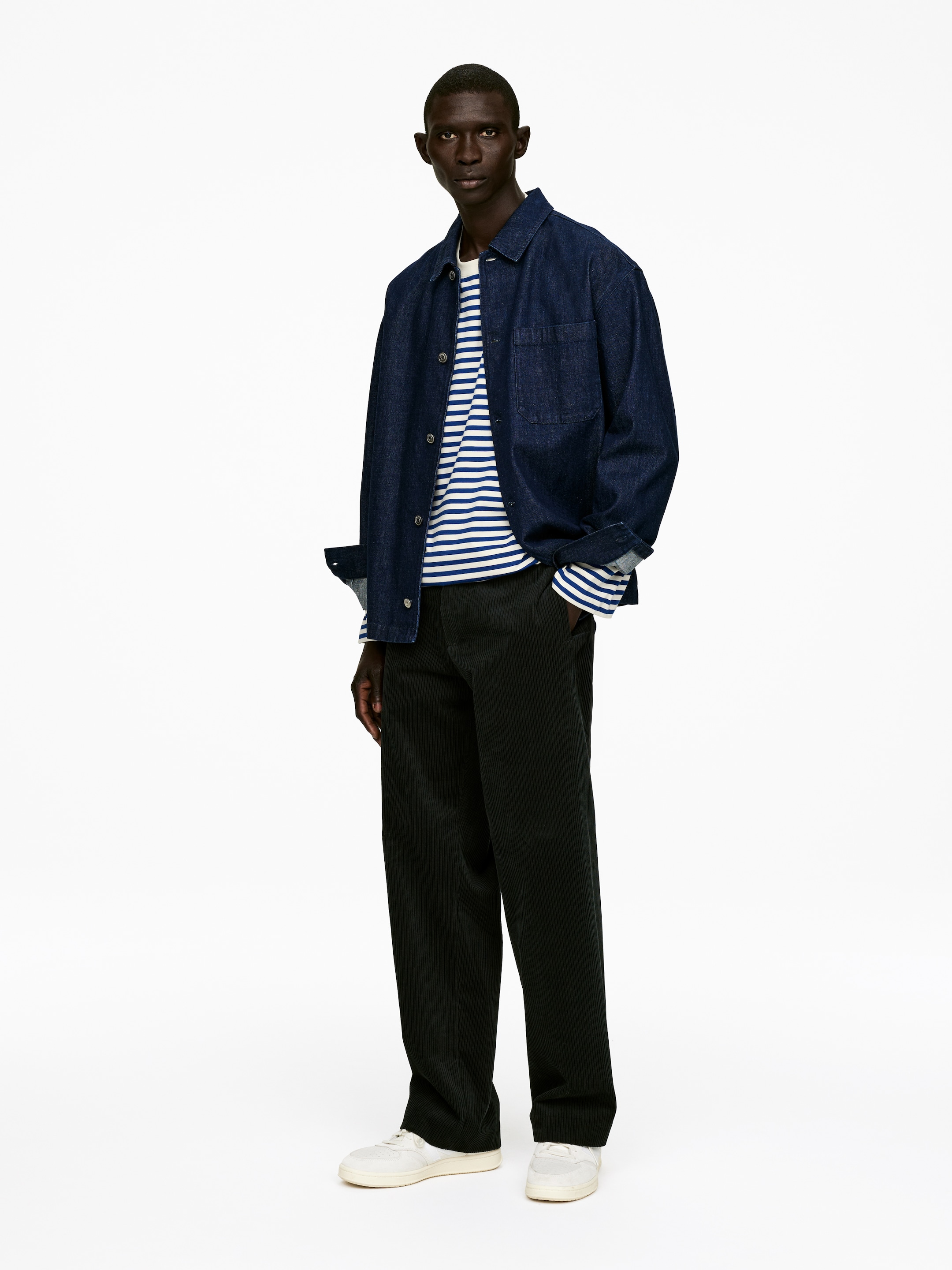 Visualizza immagine più grande: Una overshirt in denim blu scuro viene indossata aperta, le sue maniche sono arrotolate per rivelare una t-shirt a maniche lunghe a righe bianche e blu sotto, e sul davanti mostra una singola tasca sul petto. Pantaloni a gamba larga in velluto a coste verde scuro vengono indossati con sneakers bianche.