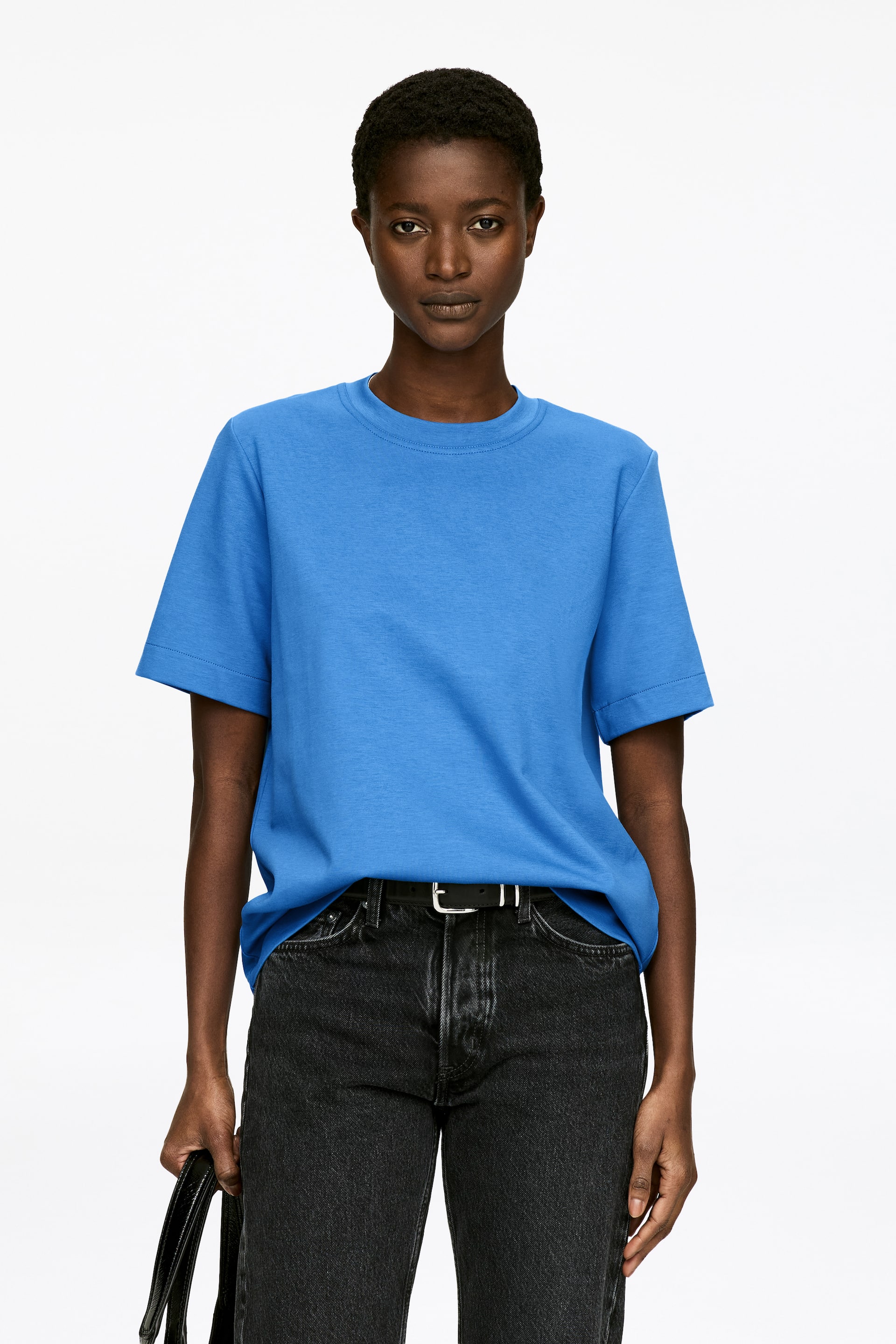 Zobacz większy obraz: Modelka ma na sobie niebieski T-shirt z okrągłym dekoltem i krótkimi rękawami. Koszulka ma regularny krój i jest częściowo włożona w czarne dżinsy, które są podtrzymywane przez czarny pasek.
