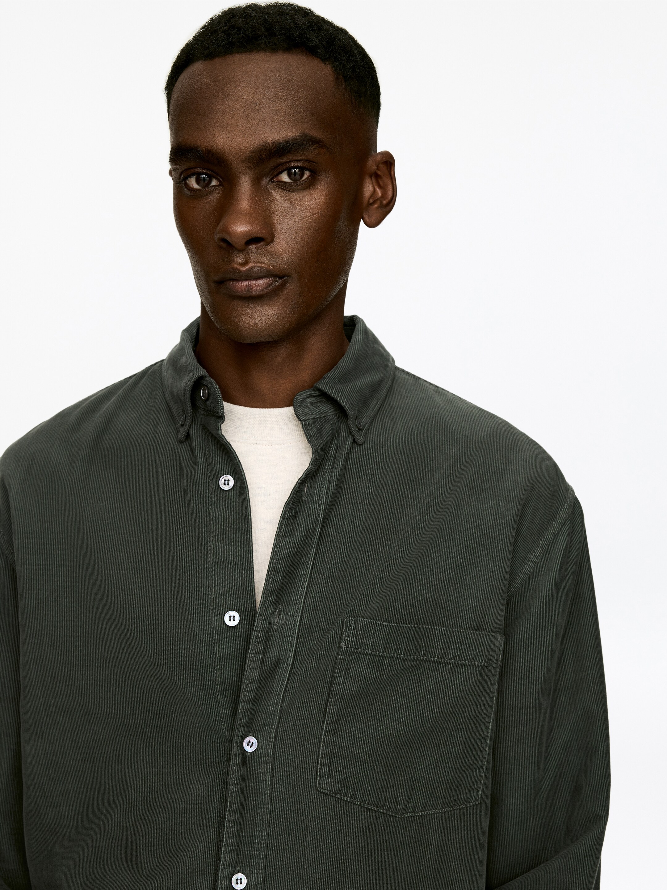 Ver imagen más grande: Una camisa de pana verde oscuro se lleva abierta sobre una camiseta interior de color claro. Presenta un cuello con botones, botones blancos en la tapeta delantera y un bolsillo de parche en el pecho izquierdo.