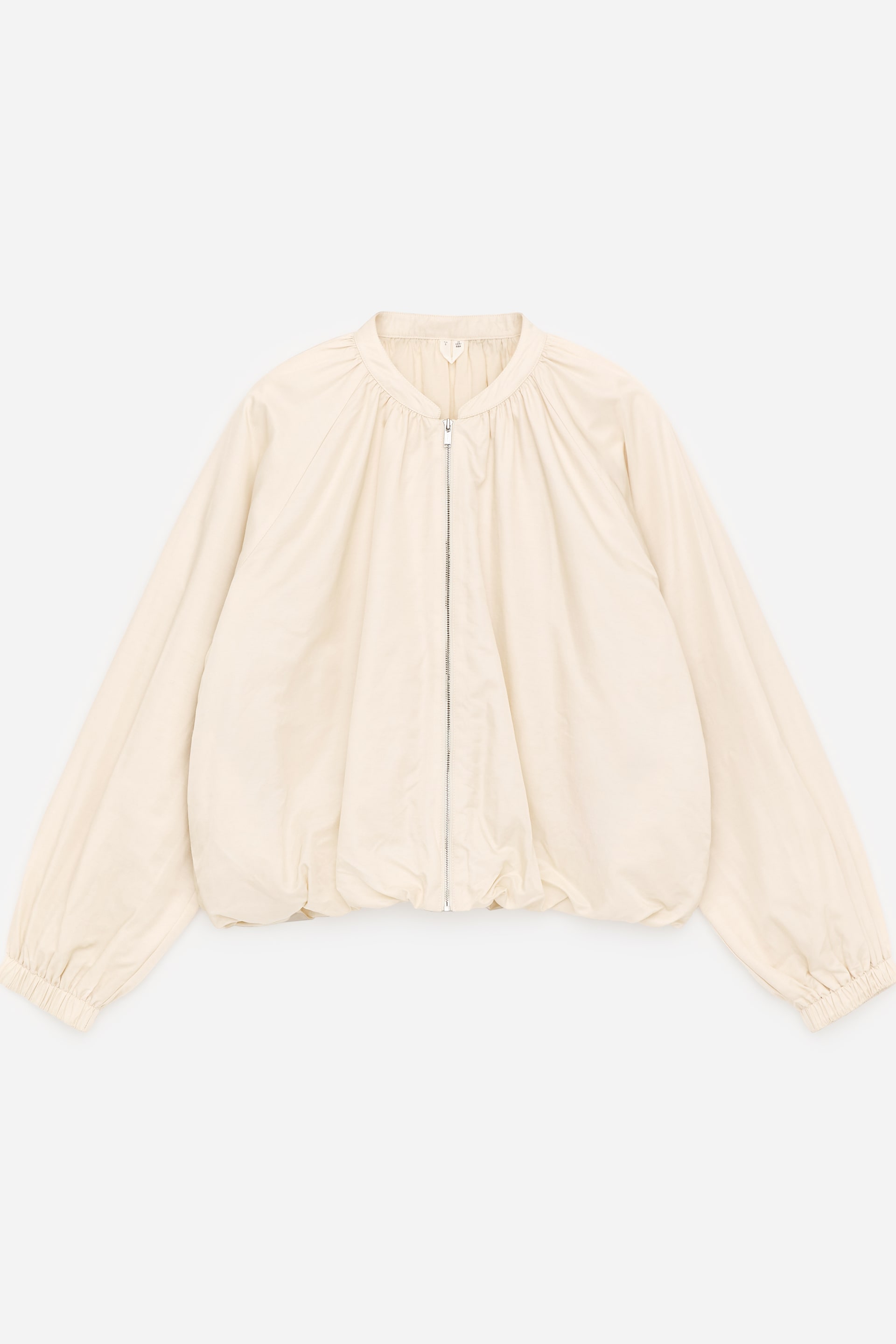 Größeres Bild ansehen: Eine leichte, cremefarbene Jacke wird von vorne gezeigt, die eine glatte, satin-nylon Qualität aufweist. Sie ist mit sanften Raffungen am Stehkragen designt, die eine dezente, ballonartige Form schaffen. Ein zentraler Frontreißverschluss verschließt die Jacke, und elastische Säume und Bündchen ziehen den unteren Saum und die Handgelenke zusammen.