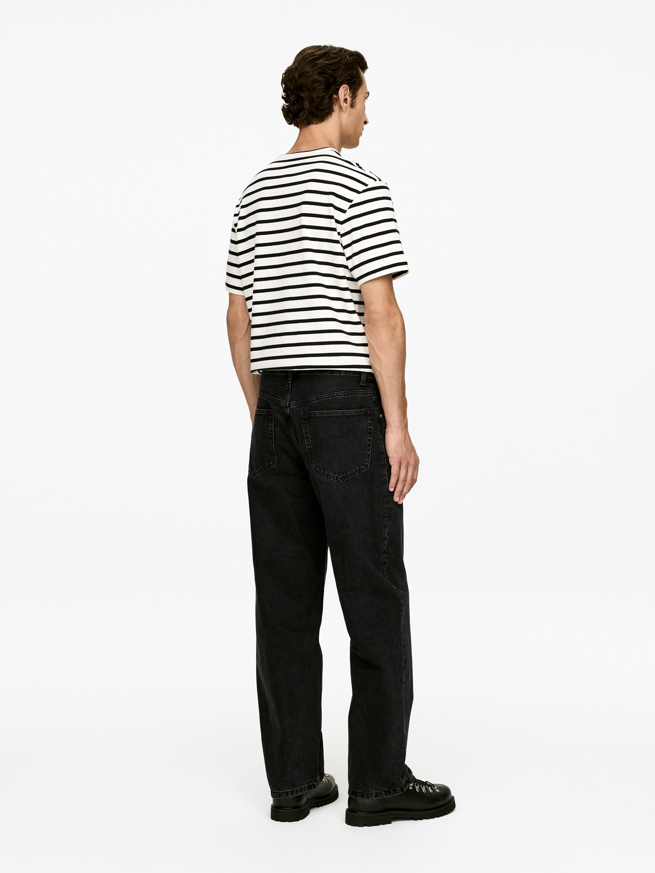 Agrandir l'image: Un t-shirt blanc à manches courtes avec des rayures horizontales noires est porté, présentant une ligne d'ourlet légèrement raccourcie. Il est associé à un jean ample de couleur sombre et des bottes noires à semelles épaisses, tous présentés de dos avec une vue légèrement de côté.